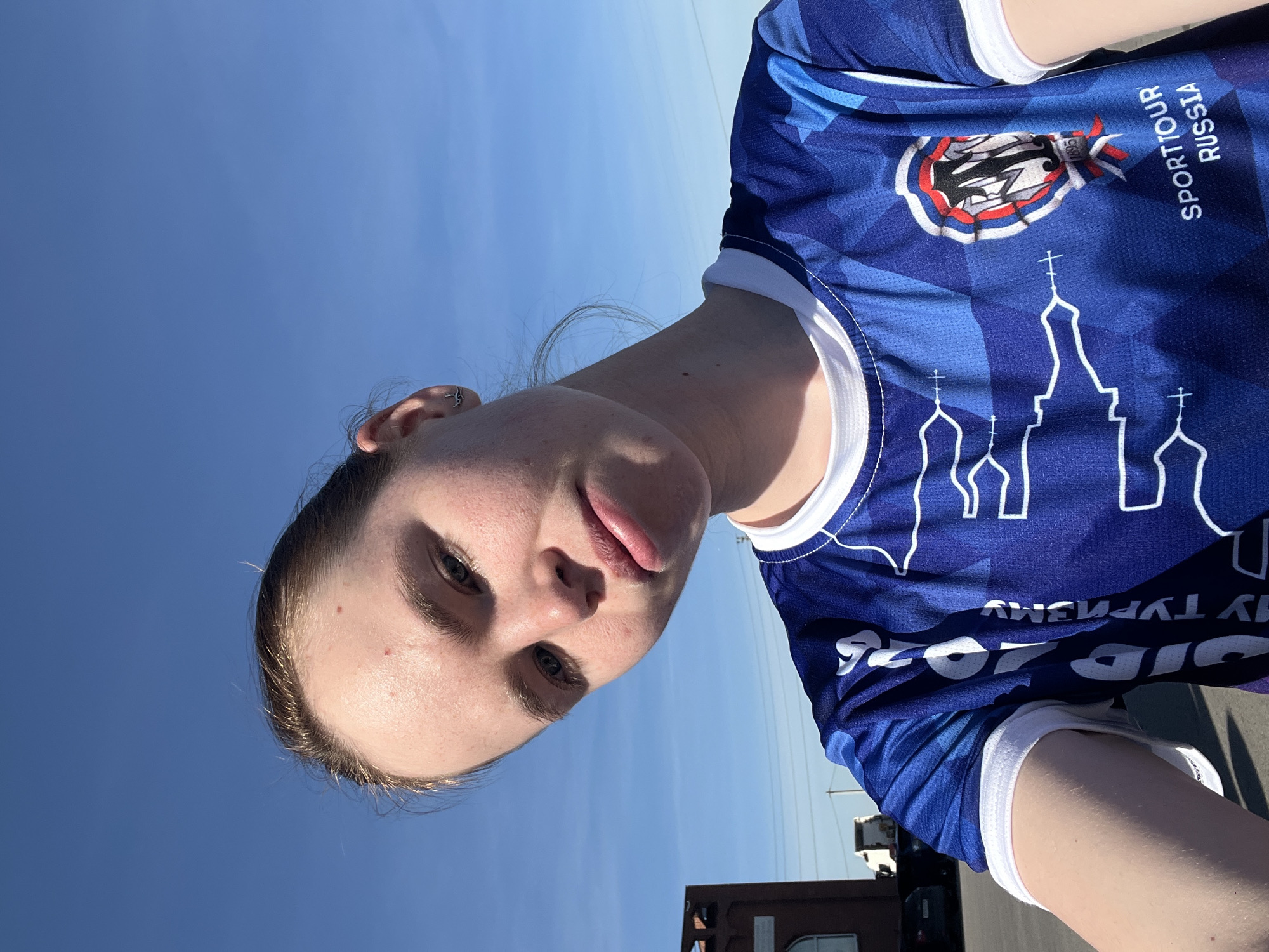 A fair-skinned person is pictured outdoors against a bright blue sky. The person wears a blue t-shirt with a white collar and white trim on the sleeves. The shirt has a pattern of darker blue geometric shapes. On the front of the shirt are white images of building silhouettes that resemble orthodox churches. There is lettering above the silhouette image that reads, "SPORTTOUR RUSSIA". Above that is a logo with a red and white design. The bottom of the shirt is only partially visible and shows reverse lettering, "SID 2026 MY TYP".
The location is Verkh-Sosnova, Russia. Other objects in the scene include a dark colored building and a black vehicle. A utility pole is visible in the distance.