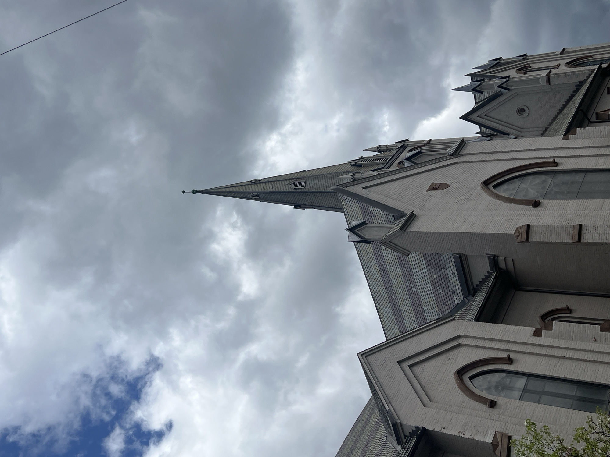 A low-angle, upward-looking photograph captures the intricate architecture of a grand, historic building, likely a church or cathedral, set against a cloudy sky. The structure, observed from a dramatic perspective, features a light-colored stone or brick facade adorned with detailed ornamentation. A prominent, slender spire with a pointed finial dominates the left side of the building, rising sharply towards the upper left of the frame. The roof sections are covered with dark, textured shingles, possibly slate, with small vents or windows visible on the spire.
Architectural details include arched windows with distinctive decorative frames, stylized projections, and intricate stonework. The sky is predominantly overcast with dense grey clouds, although a small patch of blue sky is visible towards the lower left, indicating some breaks in the cloud cover. A thin, dark cable or wire stretches diagonally across the upper left quadrant of the sky. Green foliage from a tree is just visible at the bottom right corner of the frame. On one of the building's facades, a small, rectangular plaque or inscription with faint, unreadable numbers, possibly "1893", is present. The image conveys a daytime scene under cloudy weather conditions, located in the City of Newburgh, United States of America.