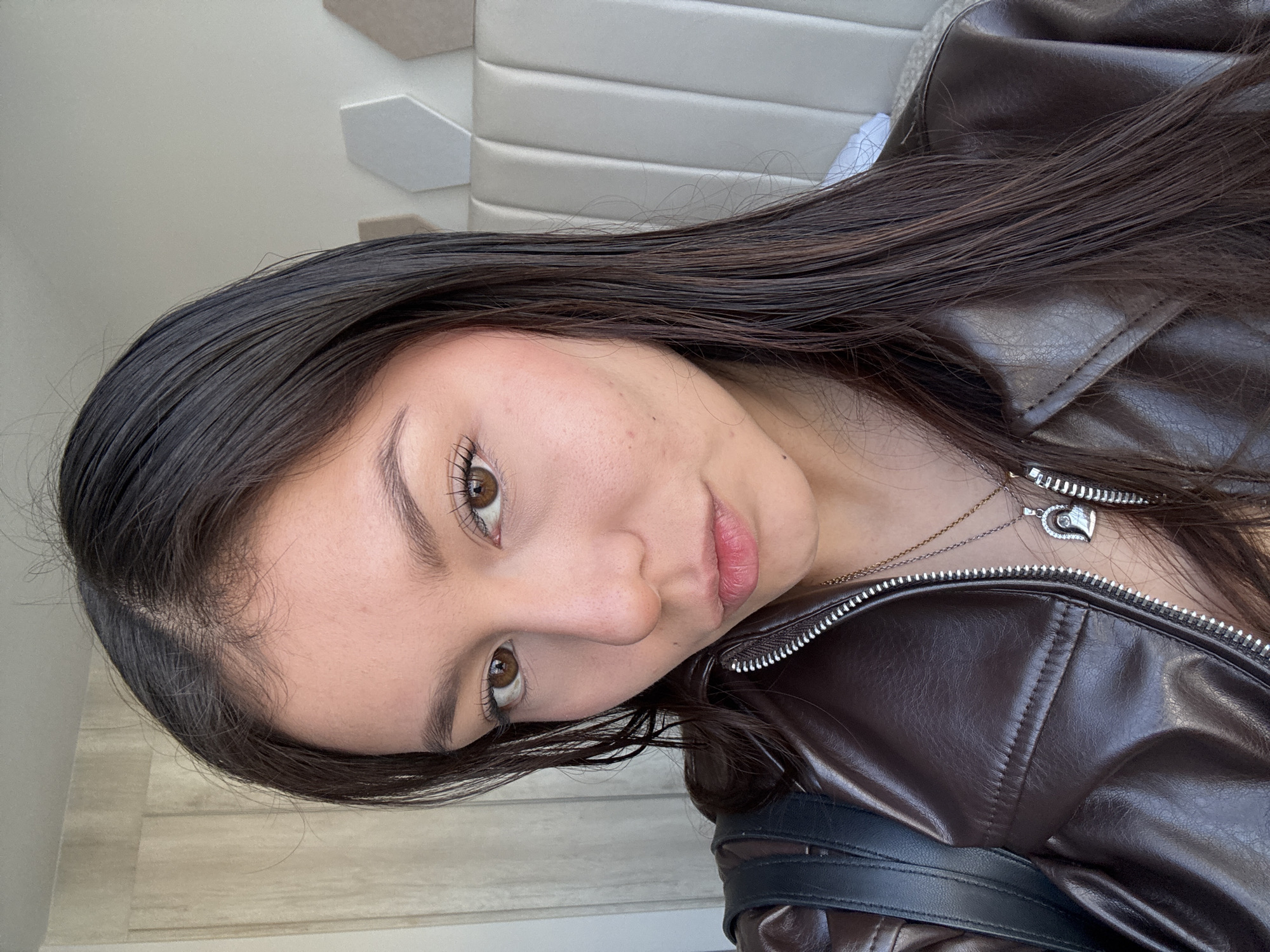 A close-up, slightly angled portrait features a young woman with long, dark brown hair and warm brown eyes, looking directly at the camera with a subtle, calm expression. Her hair is parted on the side and cascades over her shoulders. She has naturally defined eyebrows and prominent eyelashes. Her skin shows natural texture and a few small freckles on her cheeks.
She is wearing a dark brown, possibly faux leather, jacket with a prominent silver zipper running down the front. A delicate silver chain necklace with a heart-shaped pendant, featuring a smaller dark heart design inside, rests on her chest.
The background, visible behind her head and shoulders, is indoors. It features a light-colored, possibly off-white or beige, wall with a textured pattern of hexagonal or rectangular panels, which could be a headboard or decorative wall covering. On the lower left side, a light-colored wooden floor or wall panel is visible. The lighting suggests daytime. There is no visible text. The setting is in San Francisco Ocotlán (Ocotlán), Mexico.