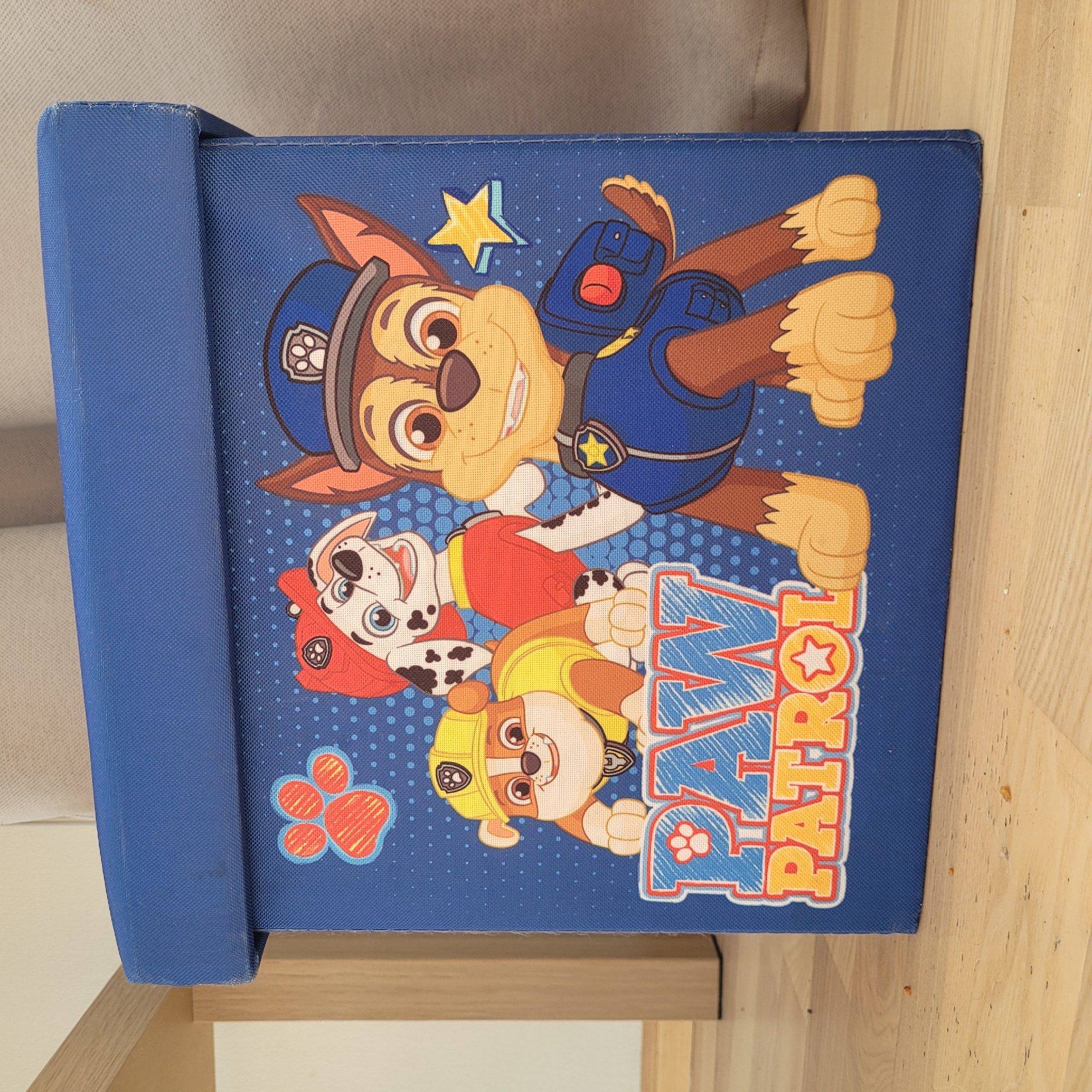 A blue toy storage box, featuring characters from the animated series "Paw Patrol," is positioned on a light-colored wooden floor. The box is decorated with illustrations of three dogs: Chase, Marshall, and Rubble, along with the "Paw Patrol" logo. A paw print graphic is visible in the upper left corner of the box. The scene is indoors, with hints of furniture and a wall in the background. The image does not provide information about its location in the city of Jūrmala, Latvia.