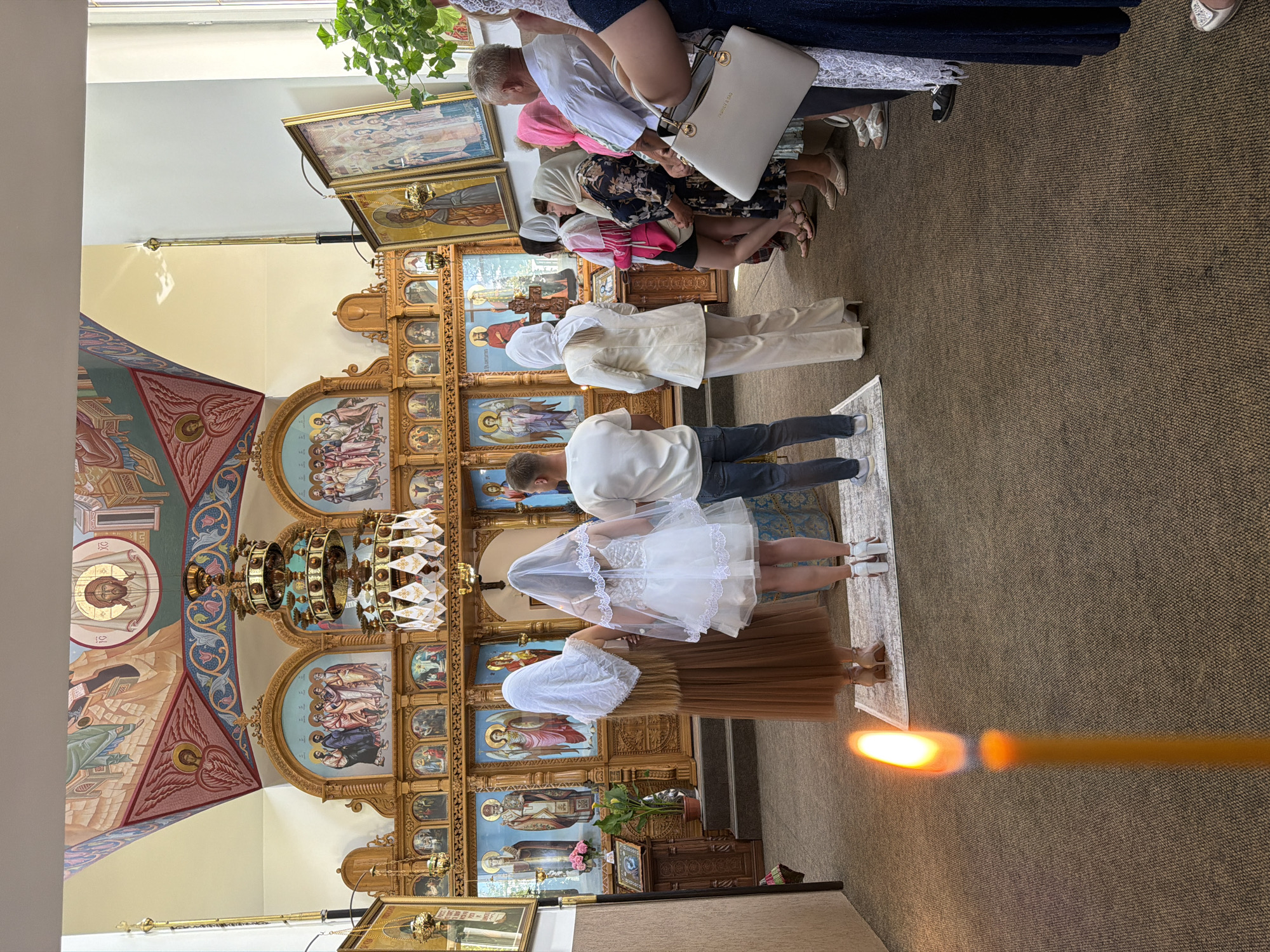 In the city of Strășeni, Moldova, an indoor Orthodox Christian religious ceremony is taking place within a church. In the foreground, a lit yellow candle stands tall, its flame casting a warm glow. The scene behind the candle reveals the rear view of several individuals, including a person in a white wedding dress and veil, possibly a bride, standing next to another person in a brown dress and white headscarf. A man in a white shirt and jeans stands to their right.
The background is dominated by an ornate iconostasis, a wall of religious icons, which is a common feature in Orthodox churches. The iconostasis is richly decorated with wooden carvings and adorned with numerous painted icons depicting religious figures. Above the iconostasis, religious murals cover the walls and ceiling, featuring depictions of saints and biblical scenes. A large, multi-tiered chandelier hangs from the ceiling, adding to the grandeur of the space.
A congregation of people, mostly women wearing headscarves, are gathered, observing the ceremony. The church interior is carpeted, and the lighting appears to be a mix of natural light from the windows and artificial lighting from the chandelier and other sources. The overall atmosphere suggests a solemn and traditional religious event.