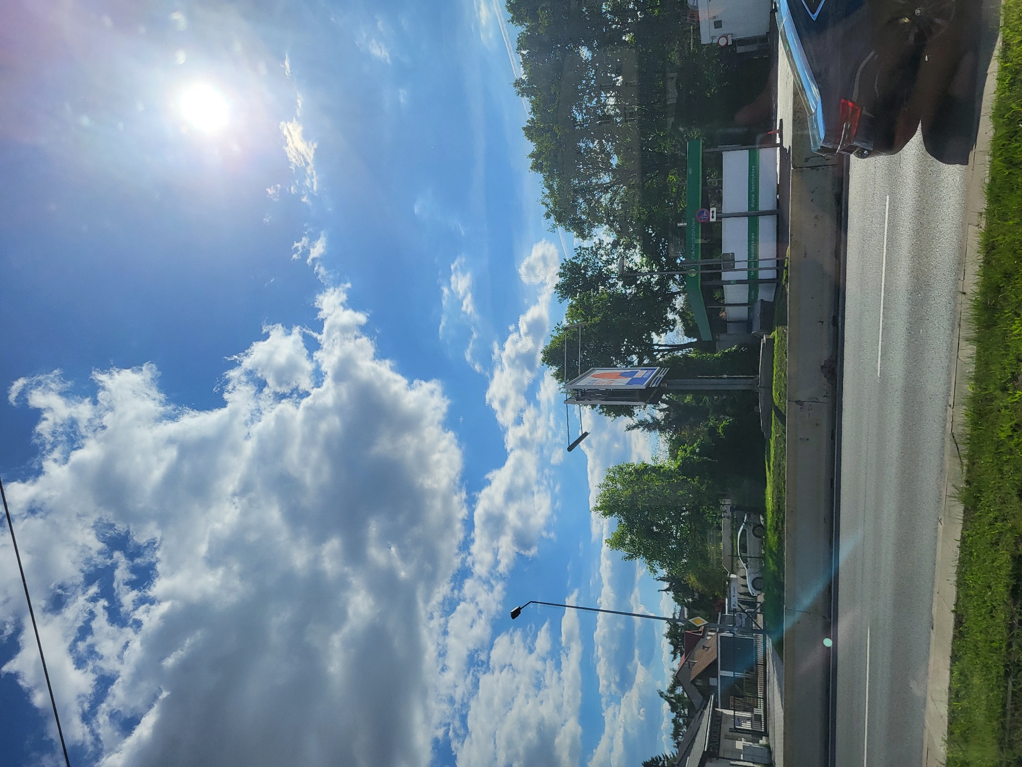 The image depicts a view from a moving car in Unknown, Unknown. The foreground includes the roadside with green grass, a gray concrete barrier, and an asphalt road with painted white lines. A black car is visible in the upper right, partially cut off by the frame. Further inland, there are trees, buildings, and street signs. The background is dominated by a bright blue sky with white, fluffy clouds and the sun shining intensely.