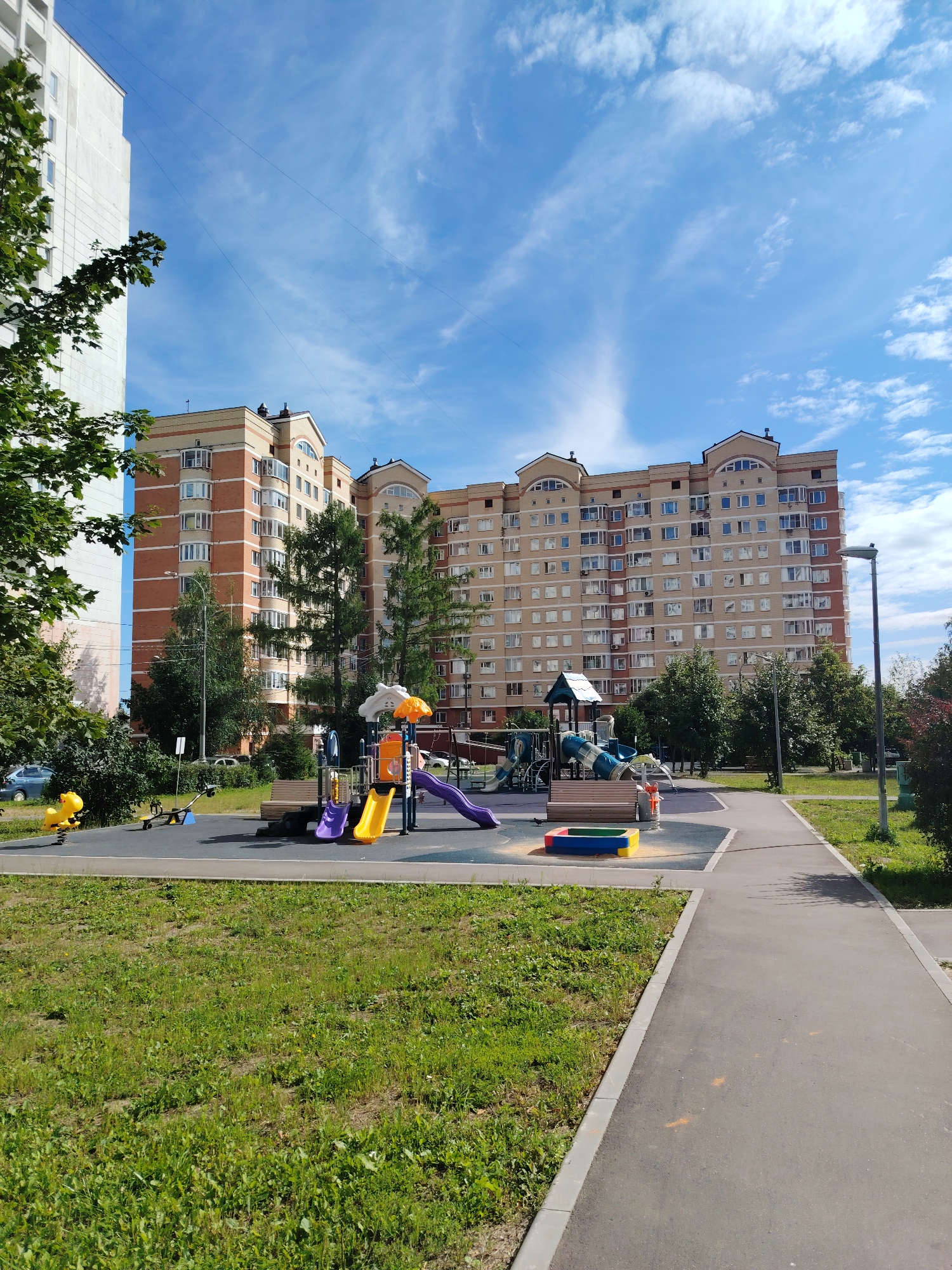 A long, multi-story apartment building with a beige facade and orange brick accents is visible in the background. The building has numerous windows and several peaked roofs. In the foreground, a children's playground is situated on a dark rubberized surface. The playground features a yellow spring rider shaped like a horse, a seesaw, a blue and green climbing structure with slides, and a colorful sandbox. A paved pathway runs alongside the playground and into the distance, bordered by a grassy area with scattered trees. A white apartment building is partially visible on the far left. The sky is blue with scattered white clouds. The location is the city of Zelenograd, Russia.
