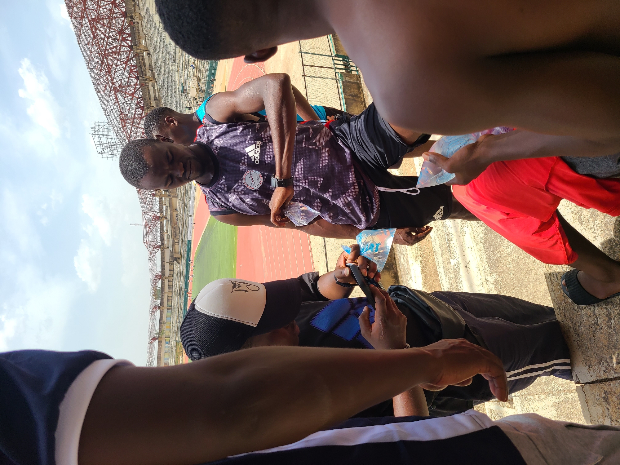 The image, rotated counter-clockwise, depicts a group of dark-skinned individuals gathered at a stadium under a bright, partly cloudy sky, characteristic of a warm daytime. The background reveals the structure of a stadium, including red metal framework, empty concrete seating, and sections of an athletic track with red and green surfaces.
Multiple young men are visible. A central figure wears a dark, patterned sleeveless athletic shirt with an Adidas logo and a circular emblem featuring an eagle and text that includes "NIGERIA." He wears a smartwatch on his left wrist and holds a clear plastic sachet, likely containing water, with "pure water" faintly visible. Behind him, another person is partially seen in a light blue top. To the right, an arm and part of a person in a red shirt are visible, also holding a similar sachet.
In the lower left quadrant, a person wearing a black and white baseball cap with a logo is focused on a smartphone. The arm and shoulder of another individual in a dark blue and white striped top are prominent in the immediate foreground.
The individuals appear to be resting or taking a break, possibly after an athletic activity given their attire and the stadium setting. They are rehydrating with sachet water, which is a common practice in Nigeria. The bright sunlight indicates either midday or early afternoon. The scene captures a casual moment among active individuals within a sports facility.