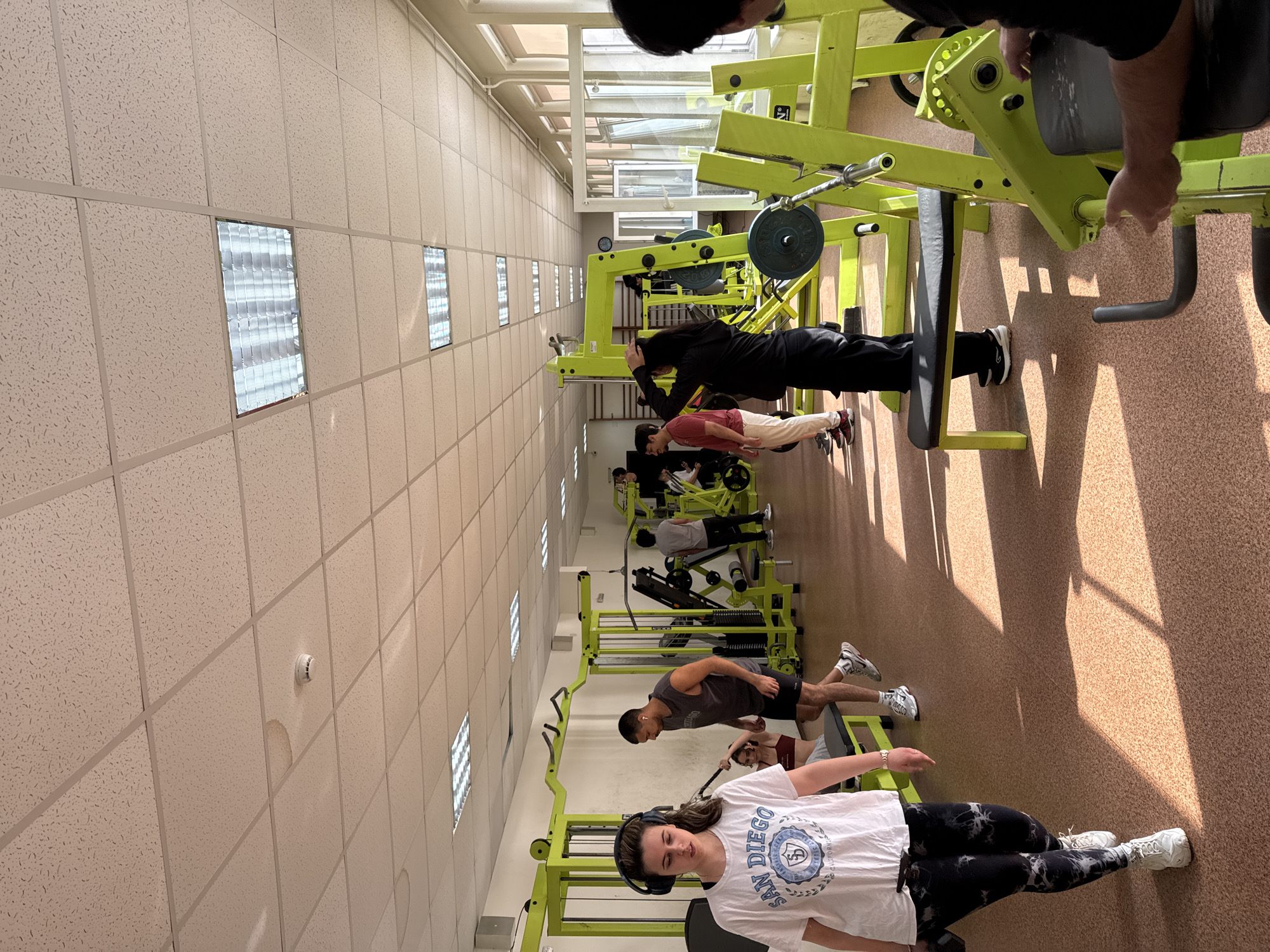 The image shows the interior of a gym, possibly in Pleven, Bulgaria, though this is not explicitly verifiable from the image. There are multiple individuals engaged in physical exercise or gym activities, primarily using weight machines.
In the foreground, a female individual wearing a white t-shirt with "San Diego" printed on it, black leggings, white sneakers, and headphones around her neck is standing, apparently not actively exercising. Further back, multiple individuals are positioned on green-colored weight machines. Some are actively using the machines, while others are standing nearby, possibly assisting or observing. The arrangement suggests a linear layout of exercise equipment along the length of the room.
The environment includes a drop ceiling with fluorescent lighting. The floor appears to be cork or a similar material. There is natural light entering the room through windows positioned higher up the wall.