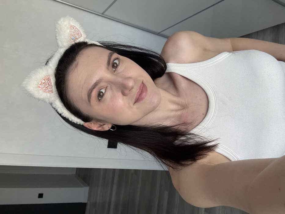 The image shows a female subject in an indoor setting, located in Kazan, Russia. The adult female has dark, shoulder-length hair and fair skin, with brown eyes and a subtle smile directed at the camera. She wears a white, ribbed tank top and small silver hoop earrings on both ears. On her head, she wears a white, fluffy headband with cat ears, the inner portions of which are adorned with pinkish-orange glitter or sequins. The subject is positioned from the chest up, seemingly holding the camera for a self-portrait. The background to the left reveals a light gray wall with a dark rectangular object (possibly a switch or panel) and a light wood-patterned floor, indicating an adjacent hallway or room. To the right, a light-colored wardrobe or closet with vertical panel lines is visible.