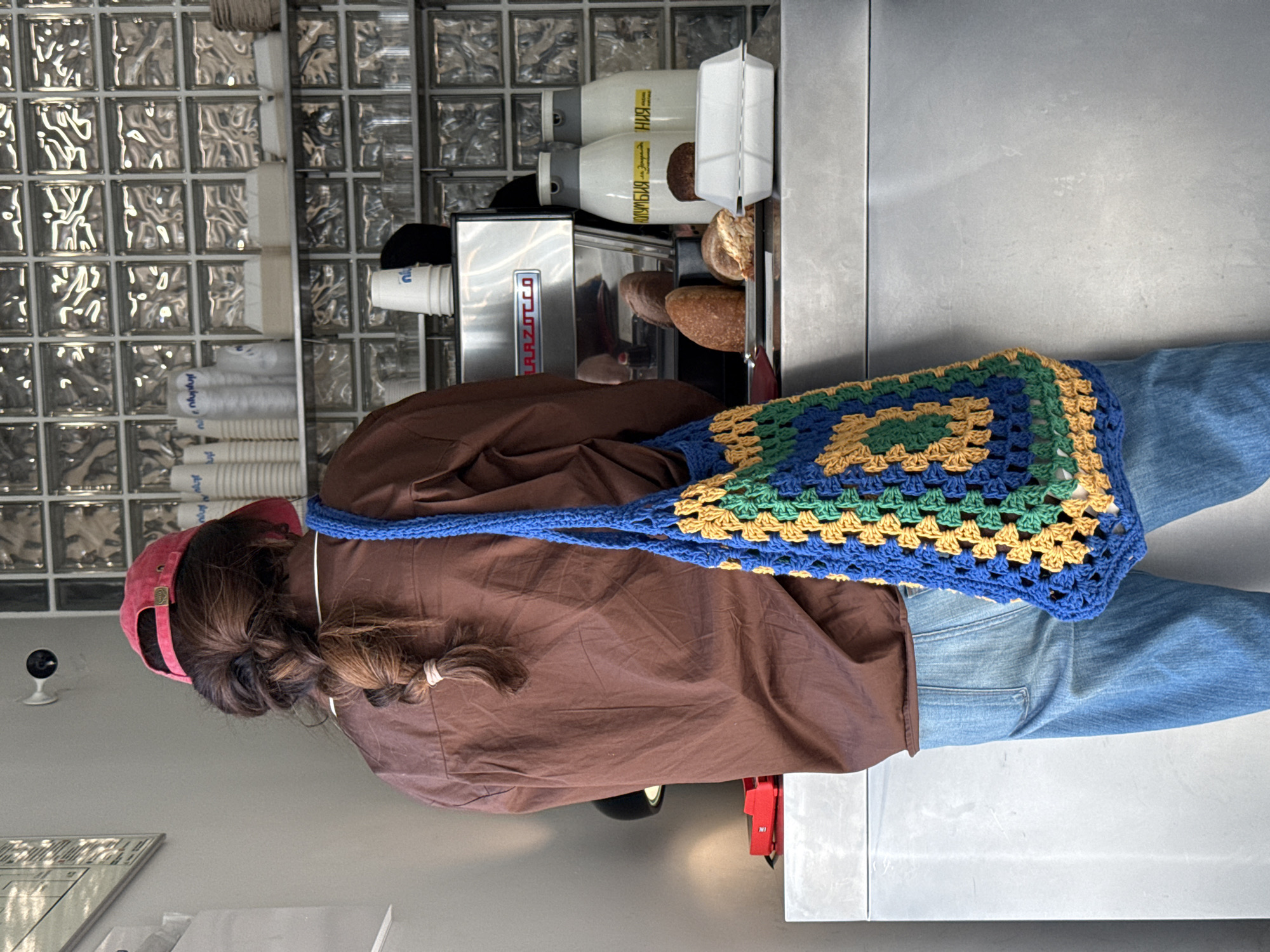 A person wearing a brown shirt, blue jeans, and a red baseball cap stands in what appears to be a cafe or bakery. They have a blue, green, and yellow crochet bag slung over their shoulder. The background features a stack of cups and a machine labeled "La Marzocco". Behind that is a wall made of glass blocks. Bread is visible near the machine. Based on the Cyrillic script on the containers, the location is likely in Moscow, Russia.
