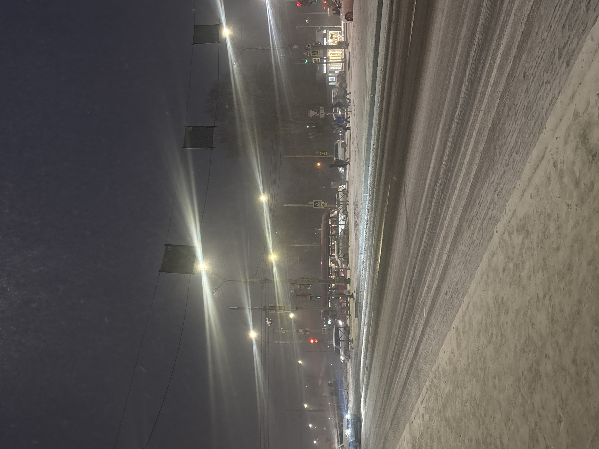 The image captures a winter night scene in Perm, Russia. The shot is taken from street level, looking toward a snow-covered road with tire tracks. Streetlights illuminate the scene, creating a hazy glow through the falling snow. Traffic lights are visible in the distance, showing red and green signals. Pedestrians are seen crossing the street at a crosswalk. Buildings and storefronts with lit windows are visible in the background. Overhead wires and suspended structures are also noticeable along the street.