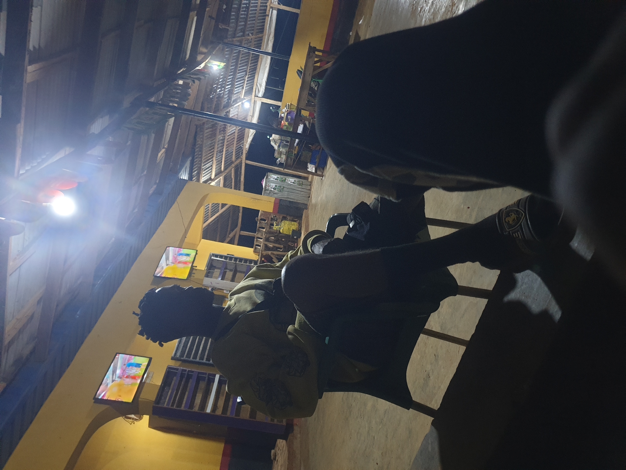 In Anilelerin, Nigeria, an outdoor covered seating area is depicted at night. A person is seated in the foreground on the left, facing away from the camera and towards two television screens mounted on a yellow wall. The televisions display images of people. The person is wearing a light-colored long-sleeved shirt and dark pants, with dark-colored sandals on their feet. A dark bag is draped over the back of the chair. The ceiling of the structure is made of corrugated metal supported by wooden beams. In the background, there is a wooden table with items on it, and shelving units against the yellow wall. The ground is paved. The overall atmosphere is dimly lit by overhead lights and the light from the television screens. Another person is partially visible in the immediate foreground, their head and shoulder in silhouette.