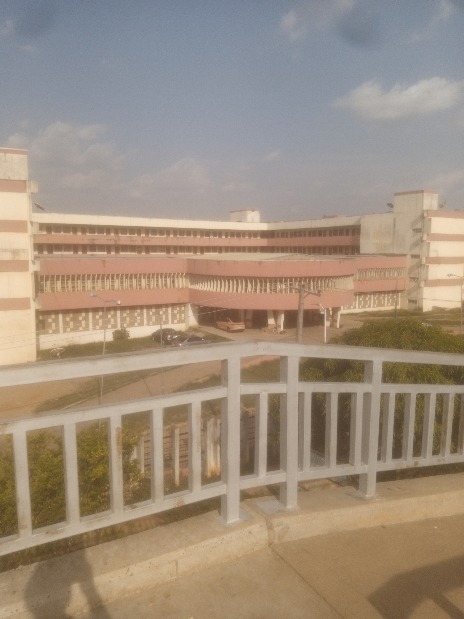 This image captures a wide shot of a large, multi-story building with a light pink facade and white trim, set against a hazy sky with scattered clouds. The building appears to be an institutional structure, possibly a hospital or university, with rows of windows on each floor. A covered walkway or entrance area is visible in the middle section of the building, supported by columns. Several cars are parked in front of the building, and there is greenery, including trees and bushes, surrounding the area.
In the foreground, a white metal railing divides the viewer's perspective from the scene beyond. The railing is made of vertical bars and horizontal supports. The ground in front of the railing is concrete, with a shadow of what appears to be a person or a tripod visible on the left side of the frame.
The lighting suggests it is daytime, likely late morning or early afternoon, given the brightness and the angle of the shadows. The overall atmosphere is somewhat subdued, possibly due to the hazy weather conditions. There are no people clearly visible in the image, and no discernible text is present. The location context provided is Unguwan Dosa, Nigeria, which places the scene in an urban or semi-urban area of that region. The building's architecture does not offer specific clues to a precise location within Unguwan Dosa without additional information.