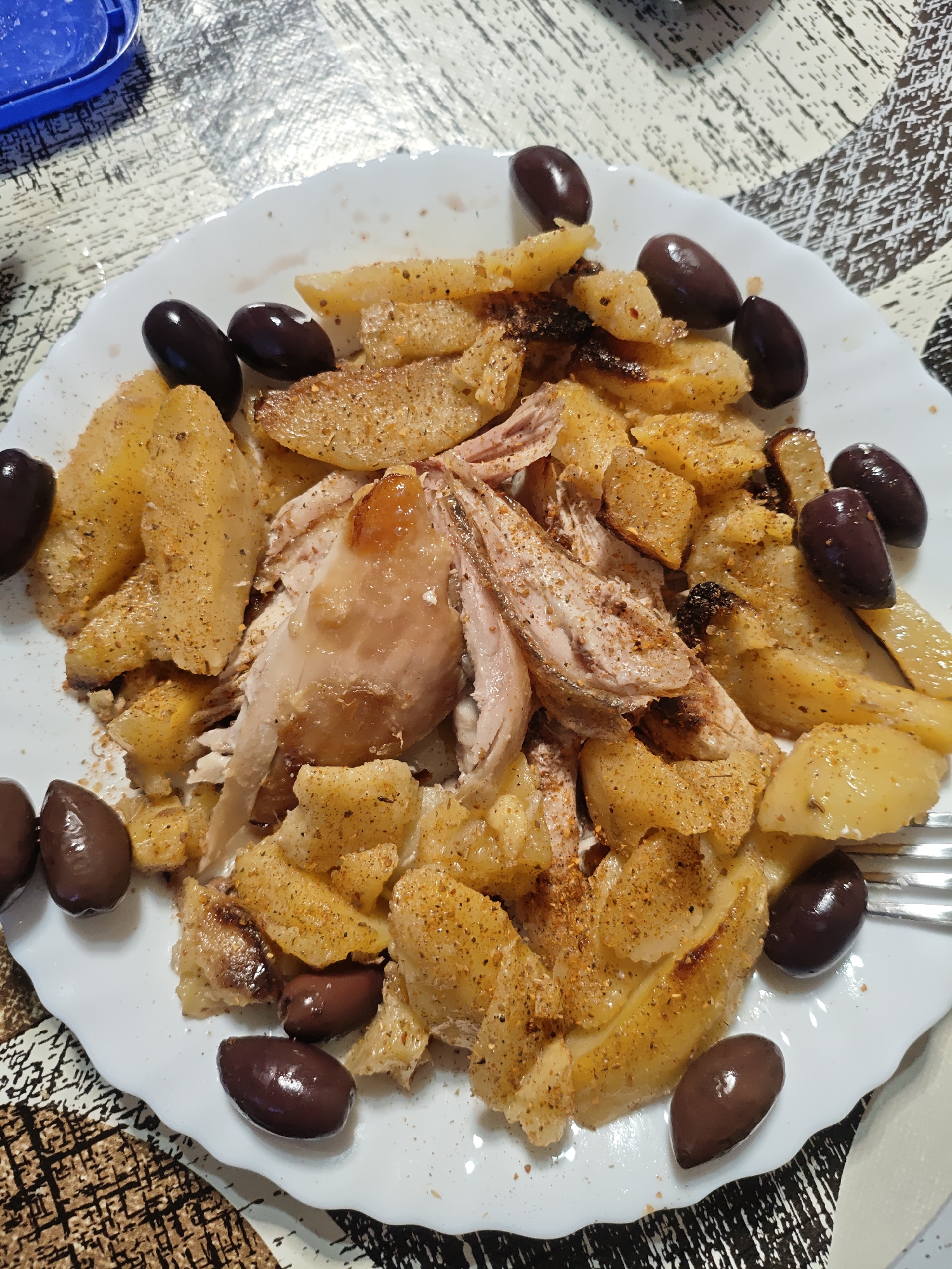 A top-down view captures a plate of food, featuring roasted chicken and potatoes, garnished with black olives. The meal is served on a white, scalloped-edge plate, which rests on a table covered with a patterned tablecloth.
The central component of the dish is shredded or pulled chicken, with some pieces appearing moist and golden-brown. It is surrounded by numerous pieces of cooked potatoes, which are cut into various shapes, some appearing like wedges or chunks. The potatoes are golden-brown and heavily seasoned with reddish-brown spices, visible as specks across their surface. Black, whole olives are arranged around the perimeter of the plate, adding a dark contrast to the lighter-colored food.
In the background, a small portion of a blue plastic container with a clear lid is visible. A silver fork is partially seen on the right side of the plate. The tablecloth features an intricate, abstract, or geometric pattern in shades of white, black, and brown, contributing to a casual, indoor dining setting. The lighting suggests an indoor environment. There is no visible text.