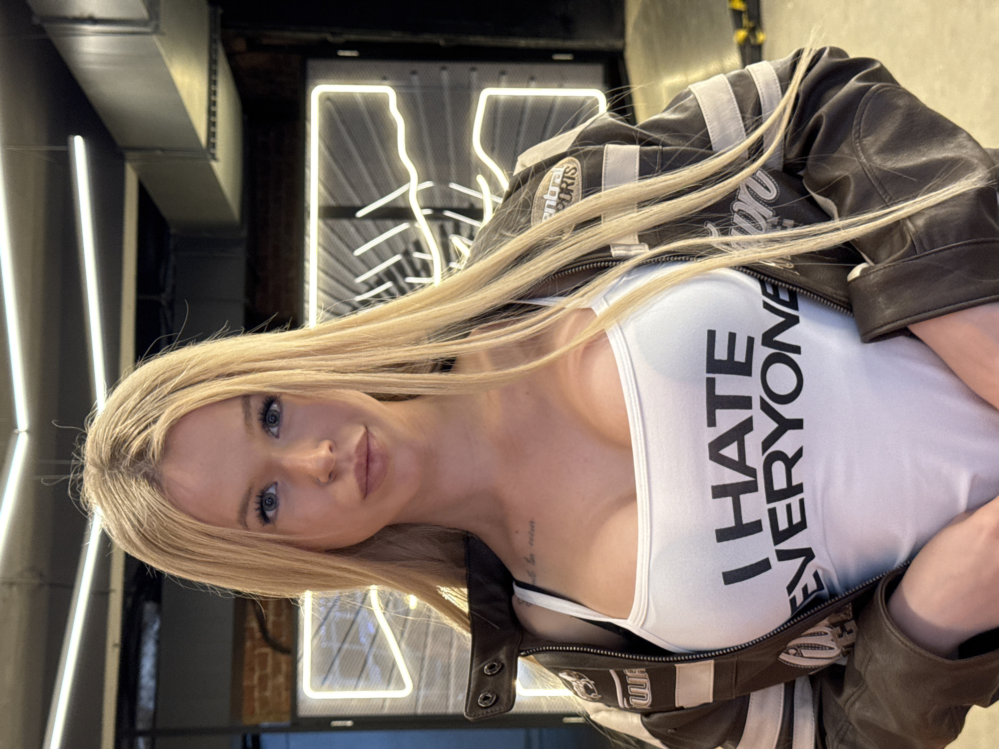 A woman with blonde hair and blue eyes is pictured in a slightly tilted, close-up shot. She is wearing a white tank top with the text "I HATE EVERYONE" printed in bold black letters across the chest. Over the tank top, she has on a brown leather jacket with white stripes on the sleeves and a patch on the left breast that reads "Central Sports". A glimpse of a black bra is visible beneath the tank top. The woman has a faint tattoo on her collarbone.
The background suggests an indoor setting with industrial-inspired decor. There are prominent neon lights in the shape of abstract figures or letters, glowing white against a dark background. Metal ductwork is visible above the neon signs. The lighting is artificial, with the neon lights casting a bright glow. The overall mood is edgy and bold, amplified by the woman's attire and the stark text on her shirt. The time of day and weather are not discernible from the image. The location context provided is Saint Petersburg, Russia.