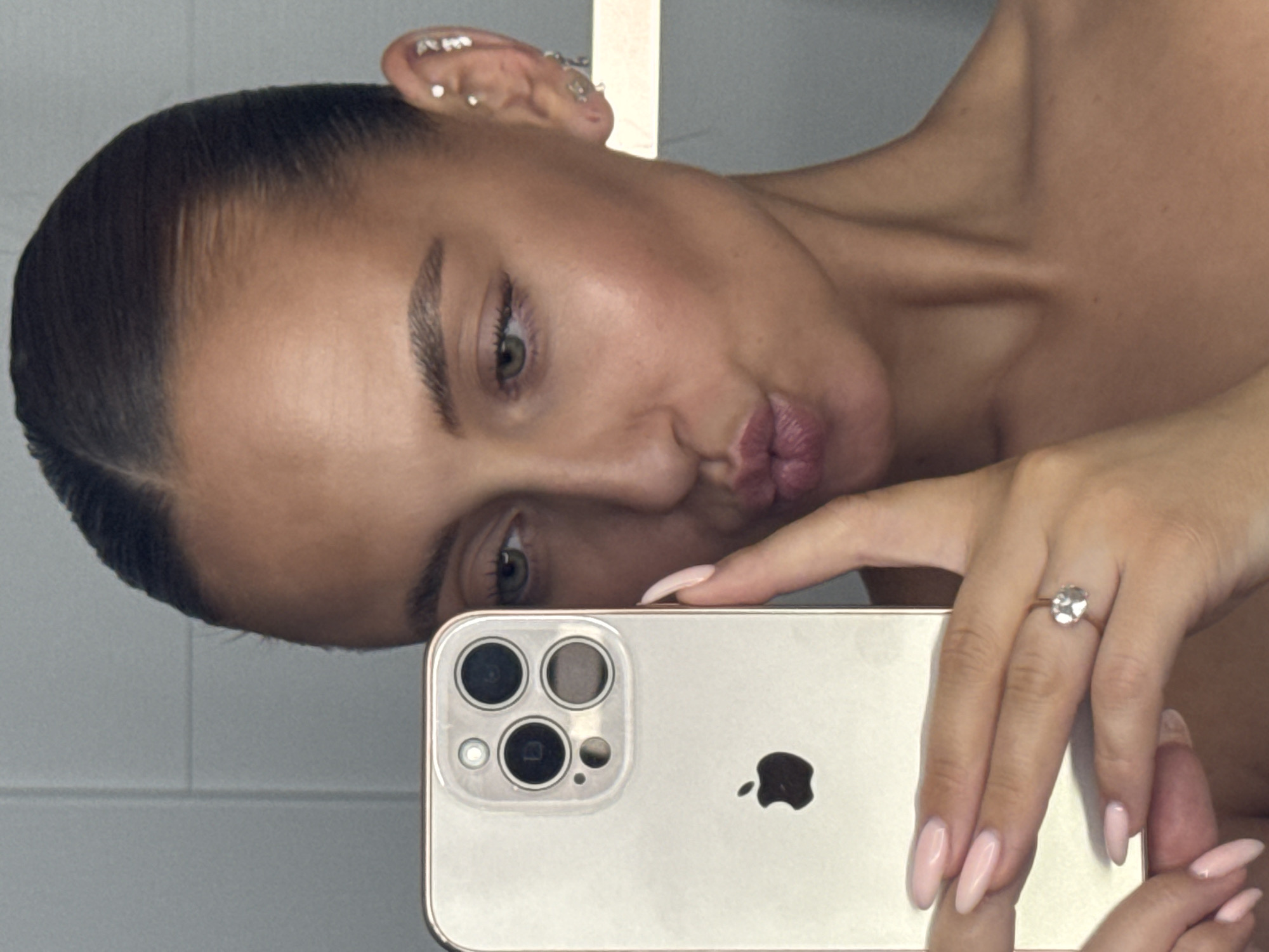 The image is a close-up, selfie-style photograph taken by a woman holding an iPhone in her left hand. She is looking at the phone's screen, with her lips puckered. Her right hand is holding the phone, partially obscuring her face. The woman has her hair pulled back tightly, and she is wearing multiple earrings in her left ear. She has a ring on her right ring finger. The background appears to be a tiled wall. The image does not contain any information or context that would allow for its location to be identified as Chaikovsky, Russia.