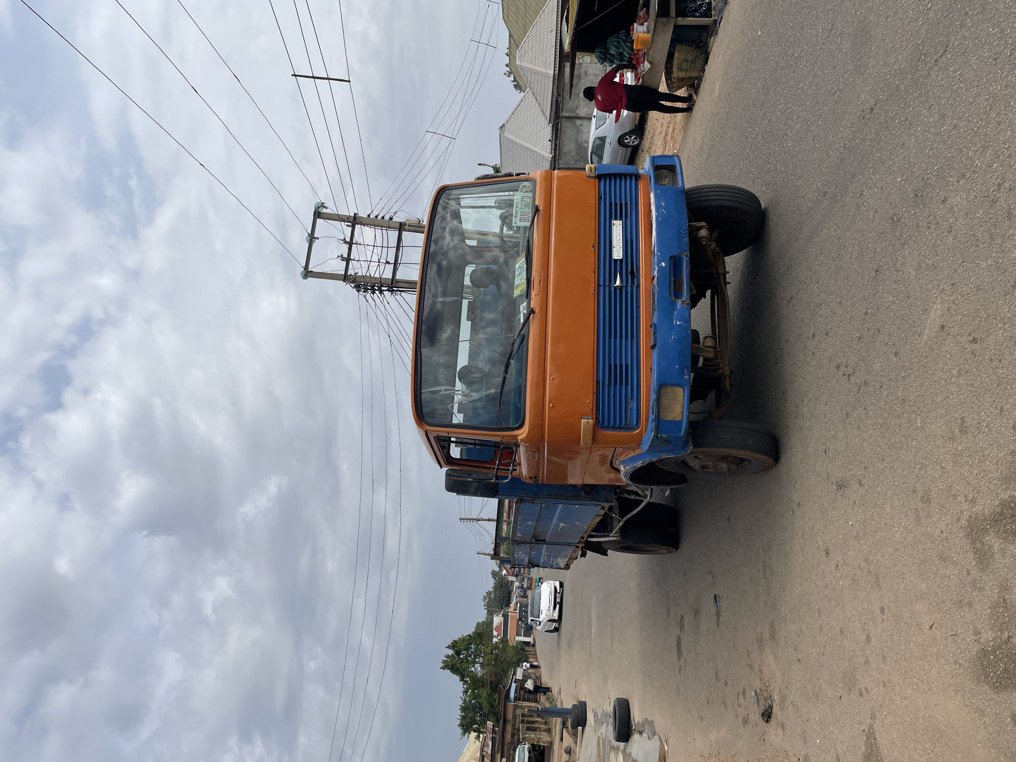 This image captures a street scene in Jos, Nigeria, characterized by daily activity under a cloudy sky. The primary subject is a large, weathered orange and blue truck, possibly a flatbed or dump truck, positioned prominently on the right side of a paved road. Its cab is orange, while the front grille and bumper are painted blue, with the word "MERCEDES" vertically visible on the grille. On the windshield, a green sticker displays "PLATE NO. KDL 279 ABJ" and "RUTV 562D."
In the background, behind the truck, a person wearing a red shirt and dark pants stands near a market stall selling goods, including watermelons. Further down the road on the left, other distant figures and a white car are visible. The general setting includes various buildings with corrugated roofs, typical of the region, and a network of power lines crisscrossing against the overcast sky, supported by a utility pole. The road itself shows signs of wear, with some loose tires or debris in the lower left foreground. The overall impression is one of a busy, functional street during daylight hours.