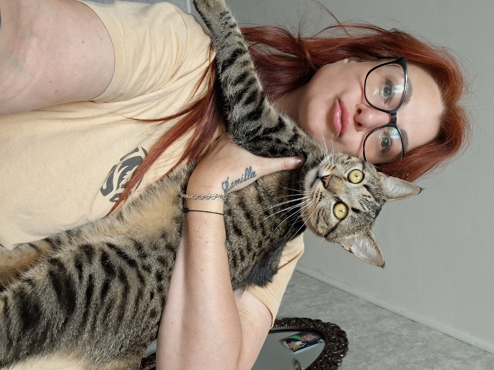 A medium shot captures a woman with auburn hair and glasses holding a tabby cat. She is wearing a tan t-shirt with a black design. The cat is looking directly at the camera with wide yellow eyes. The woman is making a kissing face. In the background, a mirror with an ornate frame reflects a child. The scene appears to be indoors. The exact location in the city of Myadzyel, Belarus, cannot be determined from the image.