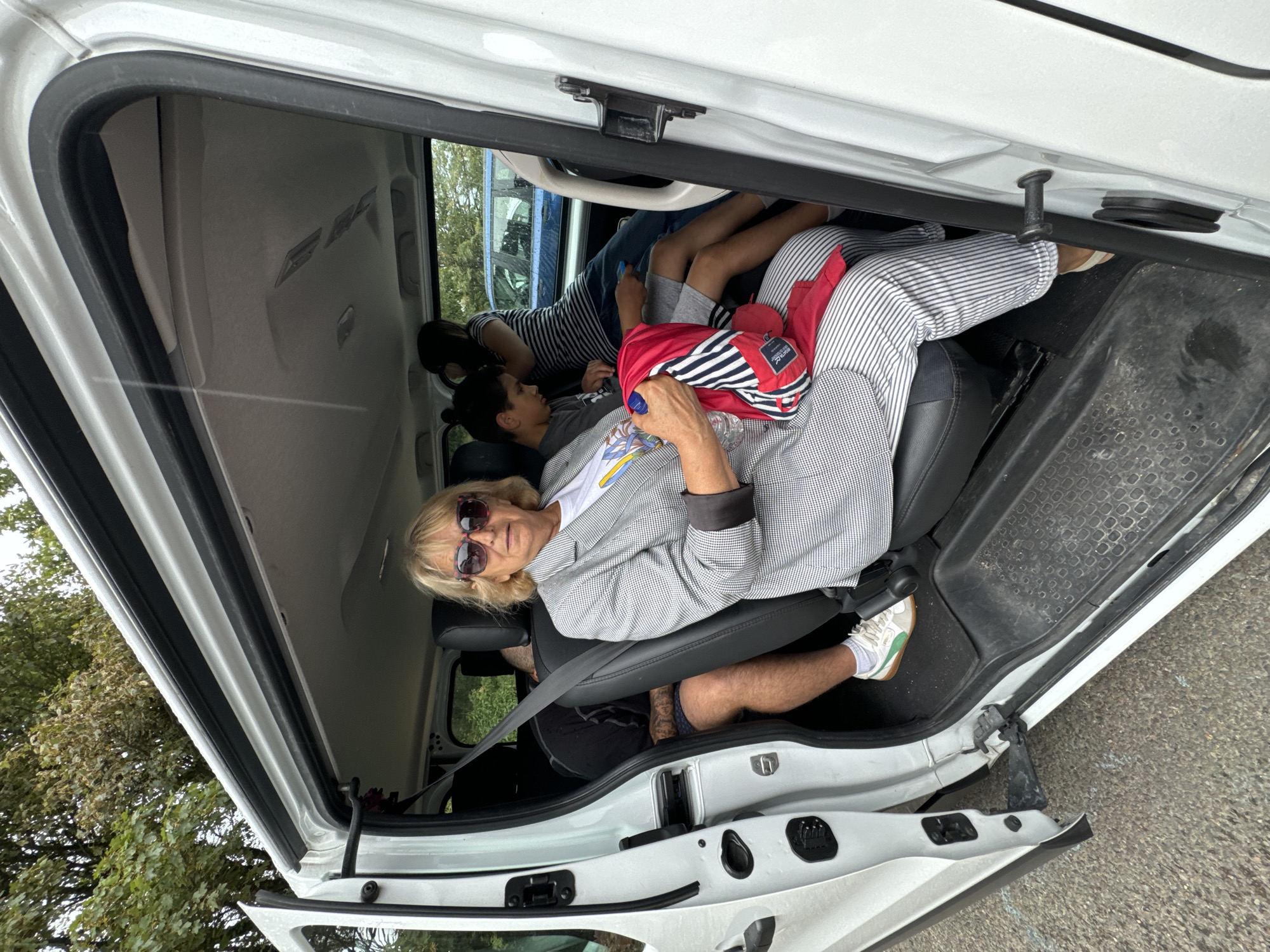 The image shows the interior of a white van with its side door open, revealing several people seated inside. In the foreground, a woman with blonde hair and sunglasses is seated in the front passenger seat, wearing a grey and white checkered blazer and striped pants. She is holding a red and white striped backpack. To her left and behind her, a man is seated, wearing a black t-shirt and patterned shorts. Behind the woman, two children are visible. One is a boy with dark hair wearing a grey t-shirt and holding a blue object. Another person, partially obscured, is seated behind the boy, wearing a striped shirt. The van's interior appears to be worn, with visible scuff marks on the floor. Outside the van, the ground is a mix of asphalt and gravel, and foliage can be seen in the background, suggesting an outdoor setting. There are no specific landmarks visible to confirm the location in Courtown, Ireland, but the overall scene depicts a casual travel situation.
