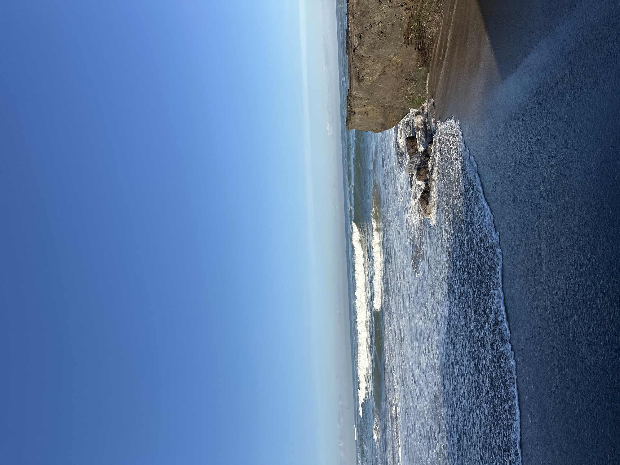 The image shows a serene ocean view with crashing waves under a clear blue sky. In the foreground, a dark sandy beach meets the foamy white surf. To the right, a large, rough-textured rock formation juts out into the water, casting a long shadow across the sand. The ocean stretches to the horizon, with several lines of waves visible, some with surfers dotting the water. The scene depicts a natural coastal landscape. Based on the characteristic waves and coastline, this scene is likely located in the vicinity of Echo Beach, a popular surfing spot in Canggu, North Kuta, Indonesia.