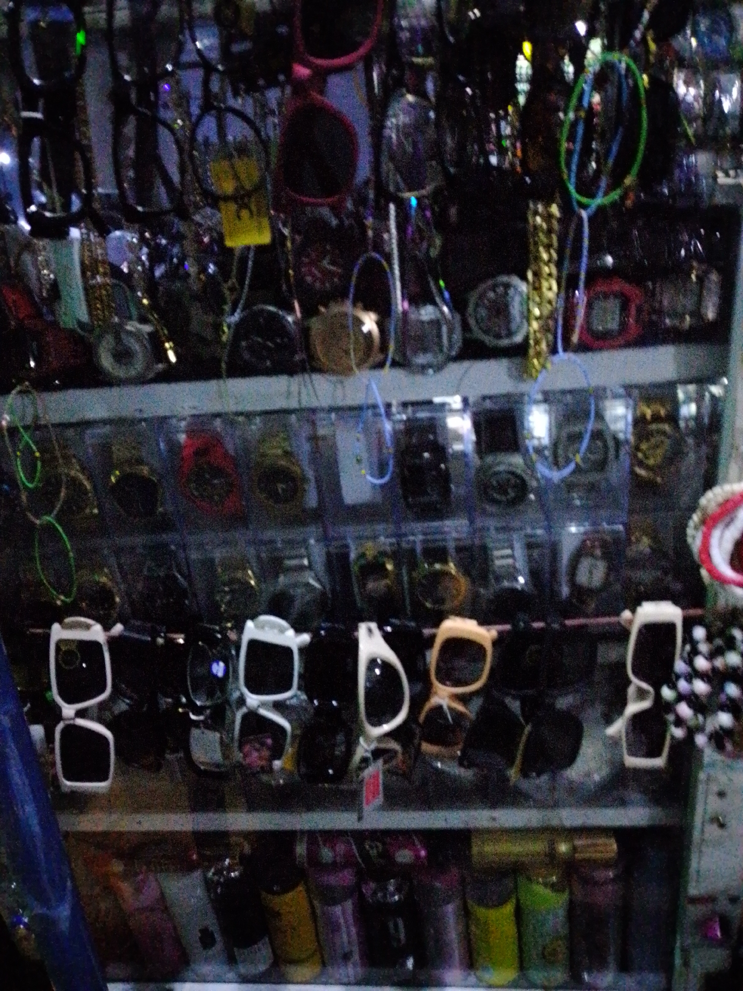This image shows a display of various items, primarily sunglasses and watches, in what appears to be a market or shop stall. The items are arranged on shelves, with many of them hanging or displayed in individual packaging. The sunglasses are of different shapes, colors, and styles, with some featuring clear lenses and others tinted lenses. Several watches are also visible, in various designs and colors, some with digital displays. In the background, there are additional rows of items, including what look like more sunglasses, watches, and possibly other accessories. The lighting is dim, and the image quality is somewhat grainy, suggesting it was taken in low light conditions, possibly at night or in an indoor setting with artificial lighting. The overall impression is of a retail environment displaying a diverse collection of fashion accessories. The context of Jalingo, Nigeria, implies this is a local marketplace where such goods are commonly sold.