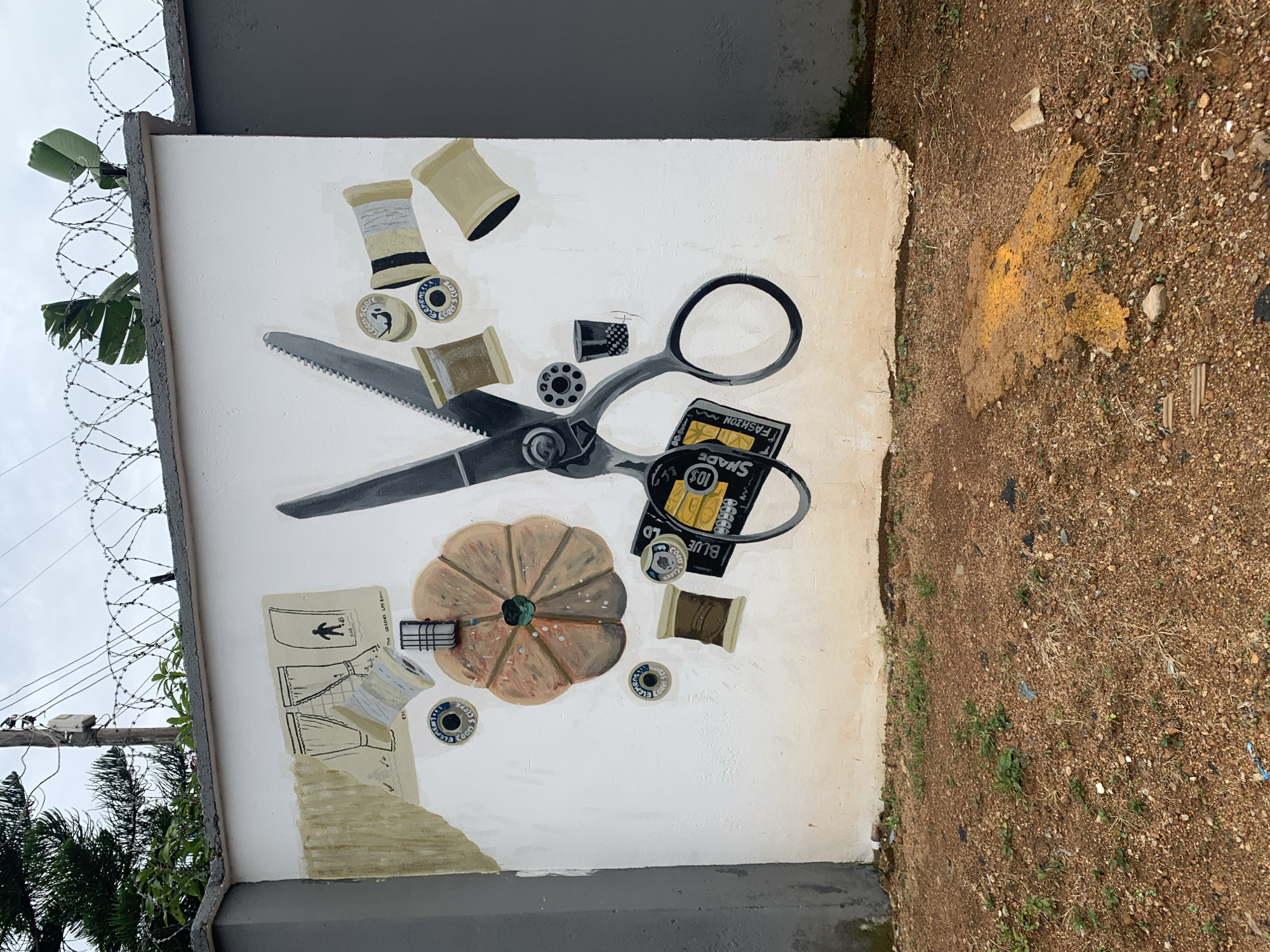 In Gura Top, Nigeria, a mural is painted on a white concrete wall. The mural depicts sewing-related items: a pair of large scissors, spools of thread, a thimble, and a needle with thread. In the upper left corner of the mural, there is a drawing of curtains and two human figures. The wall is topped with barbed wire. The ground in front of the wall is covered in reddish-brown dirt and small stones, with sparse green vegetation. There is a patch of yellow substance on the ground. The wall itself is slightly grimy and shows signs of weathering.