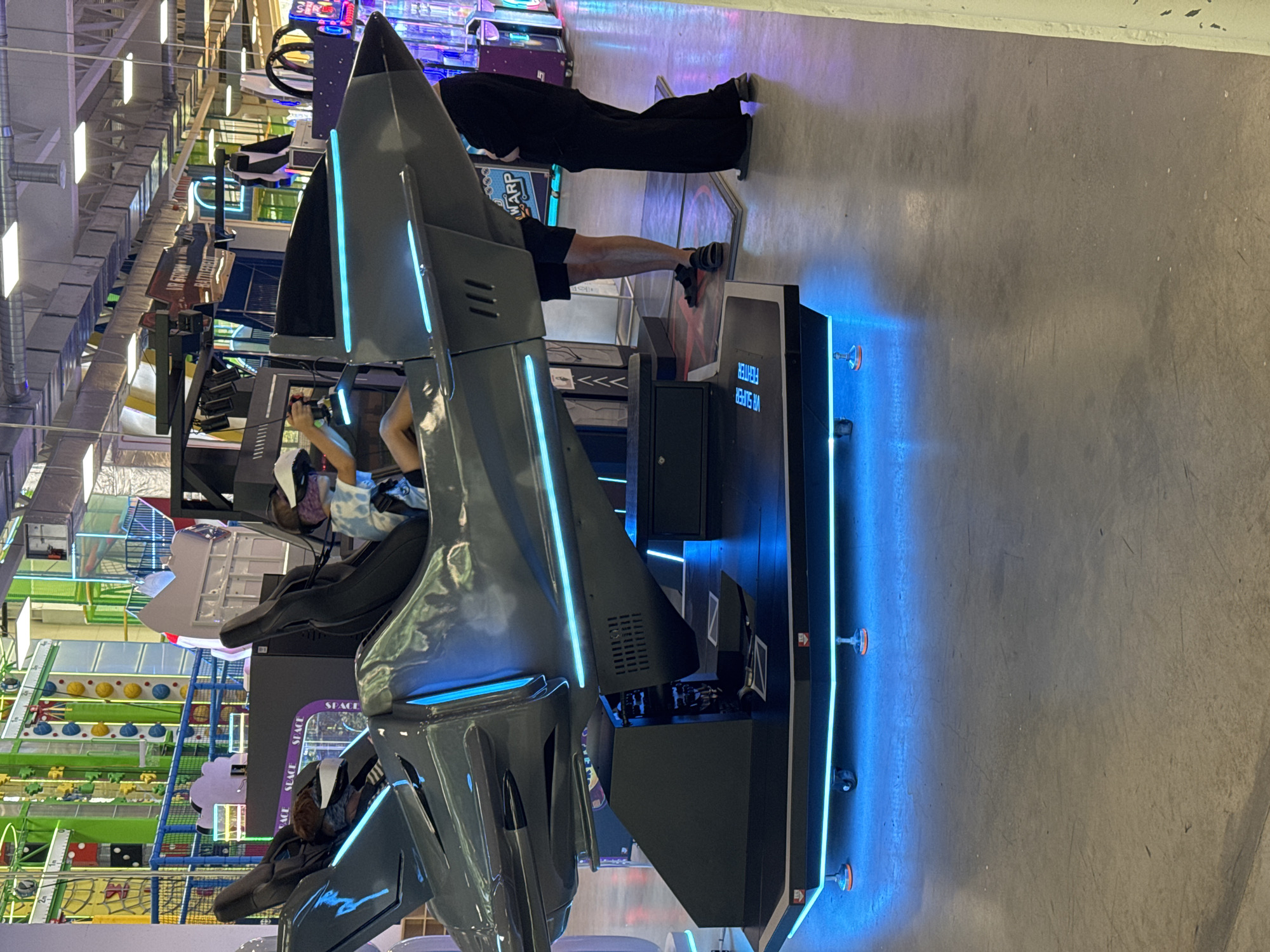 The image captures a scene at an indoor amusement or arcade facility, likely in Orlivshchyna, Ukraine, given the context. The focal point is a large, grey, jet-fighter-shaped VR simulator, illuminated with bright blue LED lights. Inside the simulator, two individuals are seated, both wearing VR headsets. One person, in the front, is actively engaged with controls, holding a joystick and wearing a tie-dye shirt. The other person is seated behind, with their head and shoulders visible.
Surrounding the simulator are various arcade games and attractions, suggested by colorful signs and machines in the background. A large climbing wall structure is visible in the far background. The floor is concrete, with some scuff marks. Two other individuals are standing near the simulator; one person, dressed in dark clothing, stands to the right, partially obscured by the simulator's structure. Another person, wearing shorts and sandals, stands to the left of the simulator. The overall environment appears to be a brightly lit, bustling entertainment venue.
