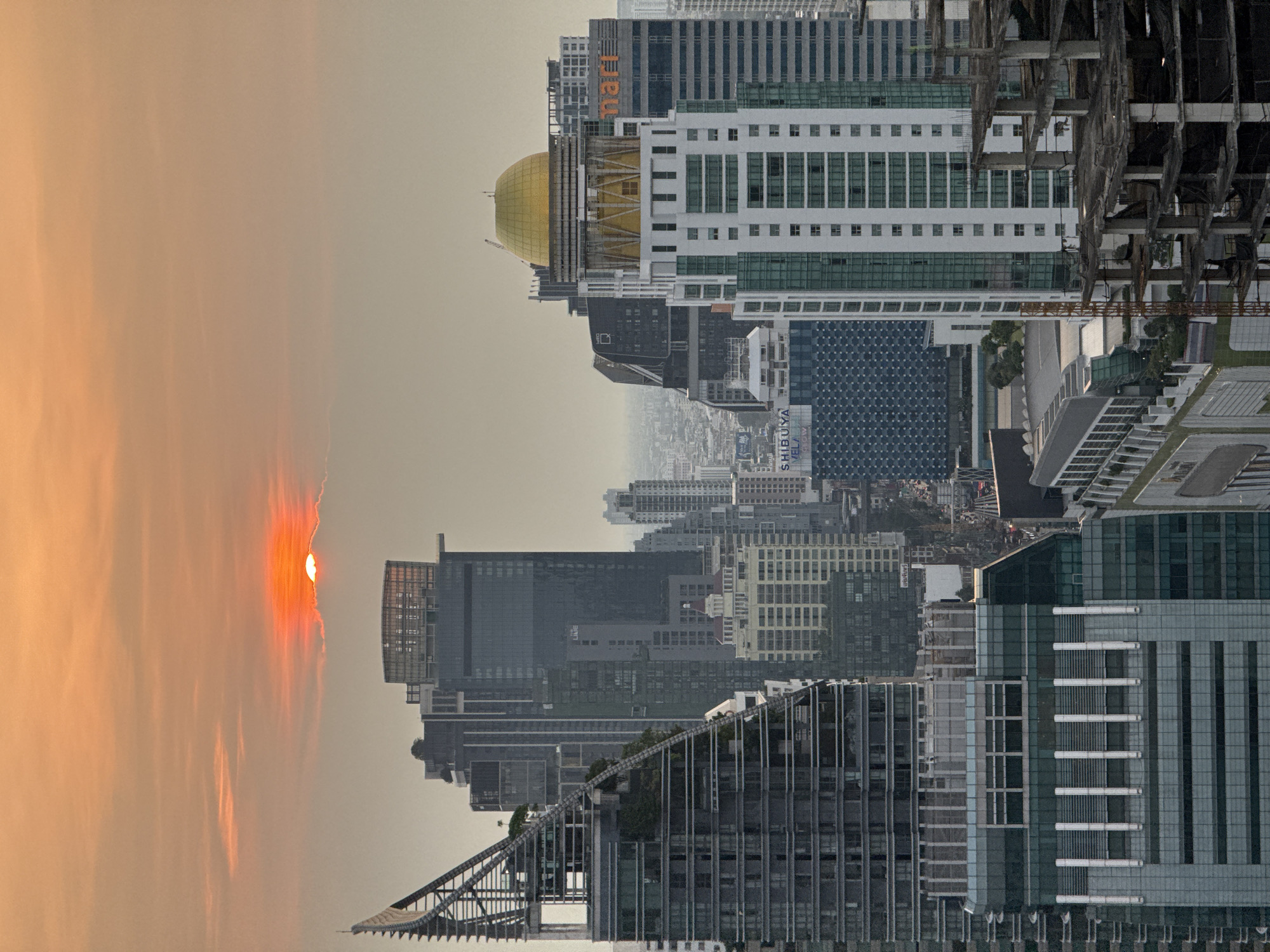 The image is an upside-down cityscape of Bangkok, Thailand at sunset. A partially obscured sun with orange and gray hues dominates the left side of the frame. Multiple skyscrapers of varying heights, materials, and architectural styles fill the lower half of the image, including the skyline of Shibuya. One prominent building features a gold dome. The sky is hazy and gray.