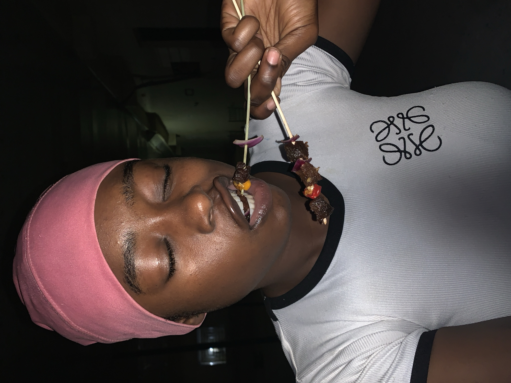 A person wearing a pink head covering and a white shirt with black trim is holding two skewers with grilled meat and vegetables close to their open mouth, with their tongue slightly extended. The background is dark, and the location is likely indoors. The Loewe logo is visible on the upper chest of the white shirt. There are no indications of New Stadium, Nigeria.
