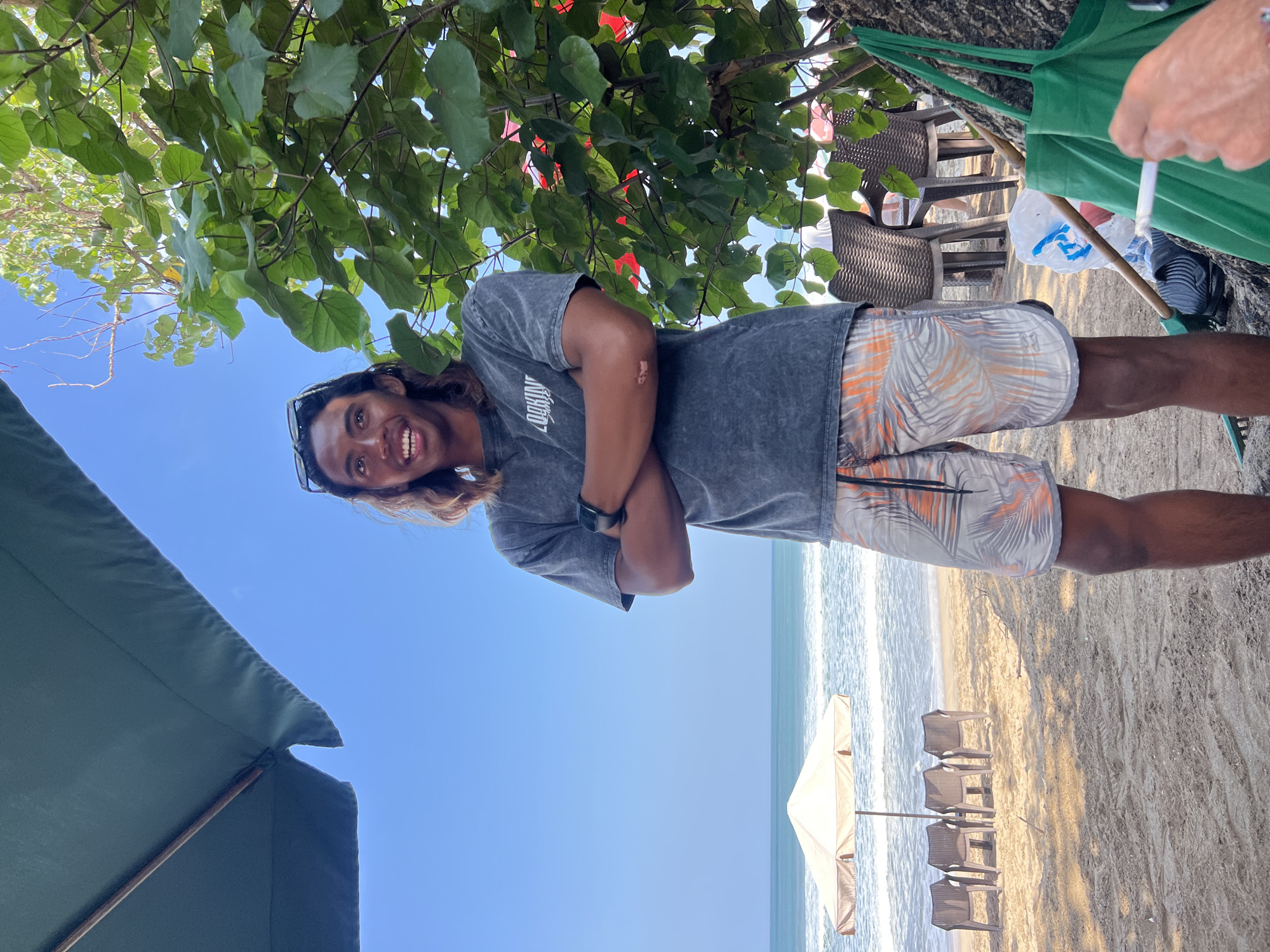 An Indonesian man with long, dark hair and sunglasses perched on his head stands on a beach in Kuta, Indonesia, with his arms crossed. He is wearing a dark gray t-shirt with white lettering and patterned shorts. The beach is sandy, with a few empty chairs and a large umbrella visible in the background, along with the ocean. A large tree with green leaves is to the right of the man, and a green tote bag hangs on its trunk. The sky is clear and blue. In the foreground, a person's hand is holding a lit cigarette.