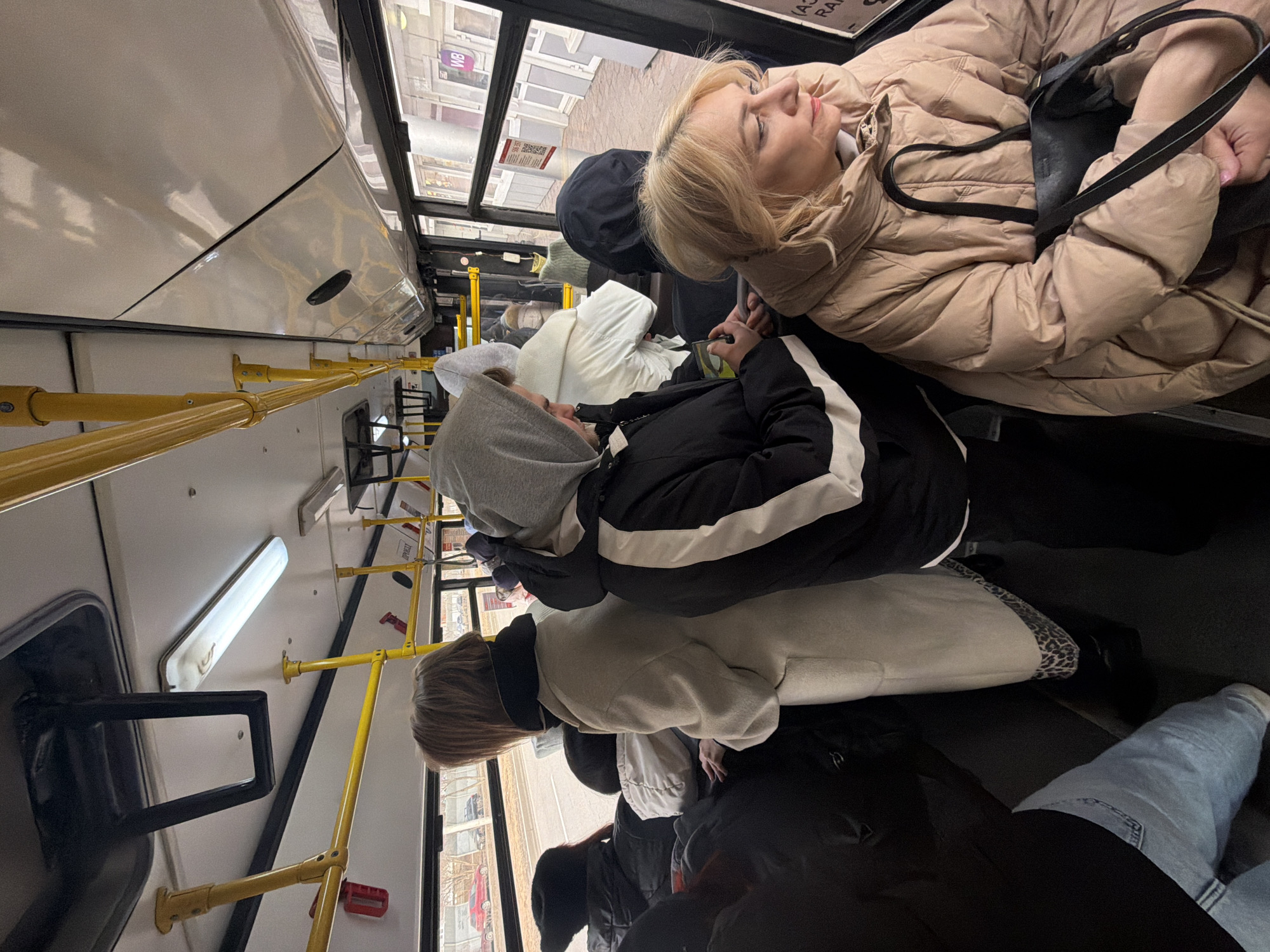 The interior of a public bus is captured in the image. Several passengers are present, each wearing winter attire indicative of a cold climate. At the front is a blonde woman wearing a beige puffer jacket and a black shoulder bag. Nearby is a person with a gray hoodie wearing a black jacket with white stripes. Further back is a person in a light-colored coat with animal-print trim. Further down the bus is a person wearing blue jeans. The bus's interior includes yellow handrails and a visible window showing buildings outside. The street sign "WB" is visible outside of the window. This suggests the bus is in Krasnoyarsk, Russia.