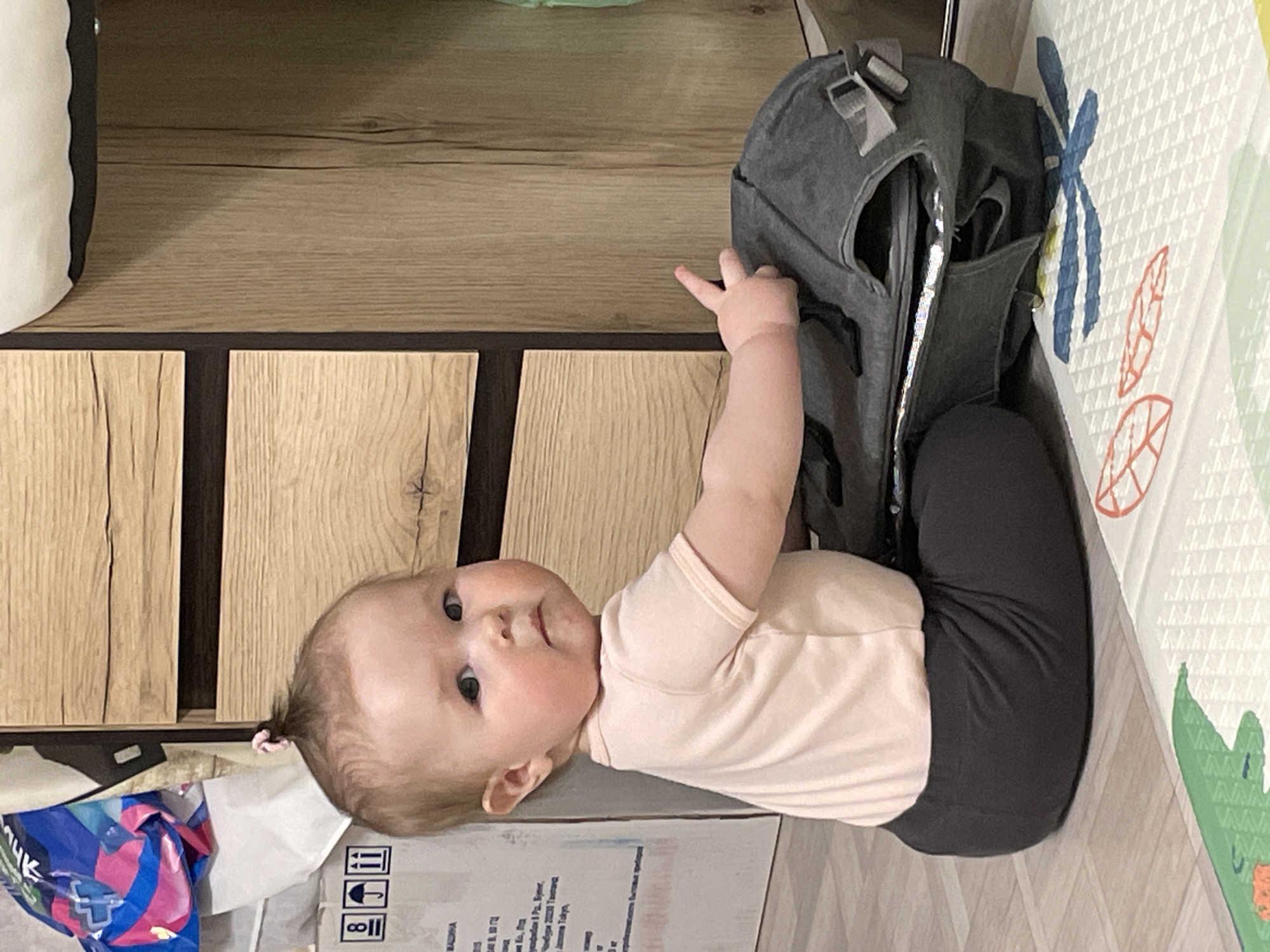 A baby is lying on a surface that appears to be cardboard or a similar material, with a gray backpack partially covering the baby's legs and torso. The baby is wearing a light peach-colored shirt and dark-colored leggings. The baby is looking at the viewer, with its right arm raised and fingers pointing toward the backpack. A cabinet with wood paneling is behind the baby. A patterned play mat is to the right of the baby and backpack. The location is the city of Chaikovsky, Russia.