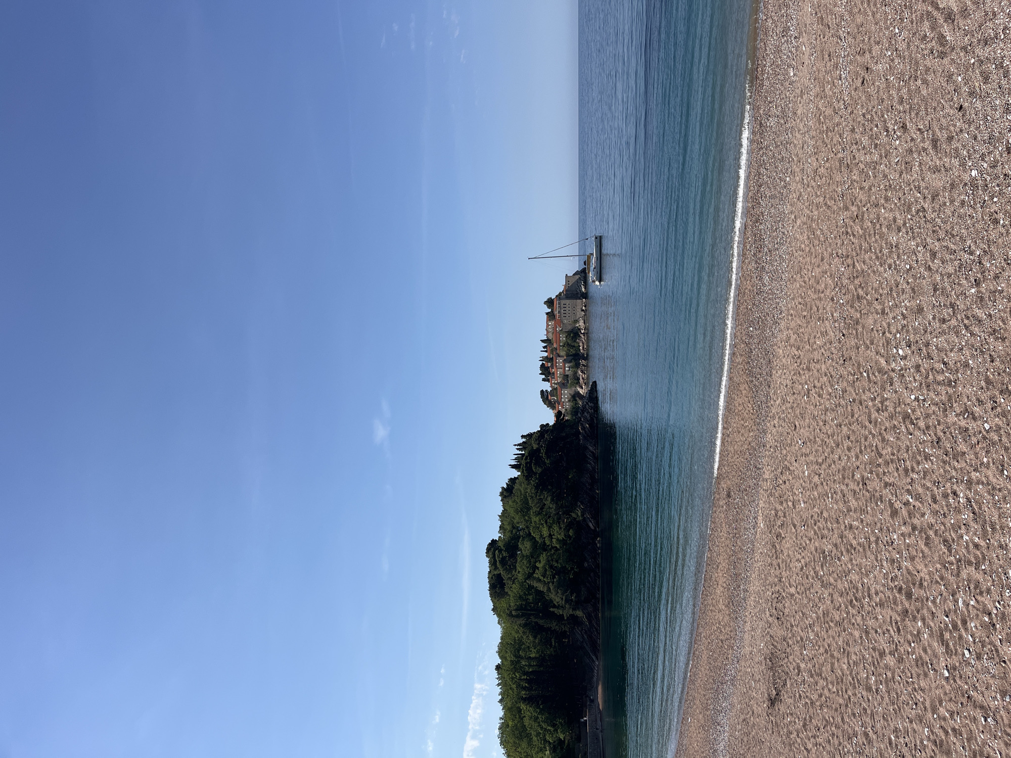 This image presents a serene coastal landscape of Sveti Stefan, Montenegro, captured on a clear, sunny day. The scene is dominated by a vast, cloudless blue sky that occupies the upper portion of the frame. Below the sky, the calm, turquoise waters of the Adriatic Sea stretch out, showing gentle ripples and reflections.
In the mid-ground, the distinctive island or peninsula of Sveti Stefan is clearly visible. The left side of this landmass is covered in dense, dark green trees and lush vegetation, while the right side features a cluster of historic, terracotta-roofed buildings built into the rock, characteristic of the iconic resort.
An elegant white sailboat with a tall mast is anchored in the tranquil waters directly in front of the Sveti Stefan island, adding a touch of nautical charm to the vista. The foreground of the image features a wide, pebbly beach where the calm sea gently meets the shore, marked by a thin line of white foam from the waves. The beach sand is light brown, speckled with smaller stones and shells.
The overall impression is one of peaceful natural beauty and historical charm. There are no people visible, and no discernible text appears in the image. The bright daylight indicates it is likely morning or afternoon, with excellent weather conditions.