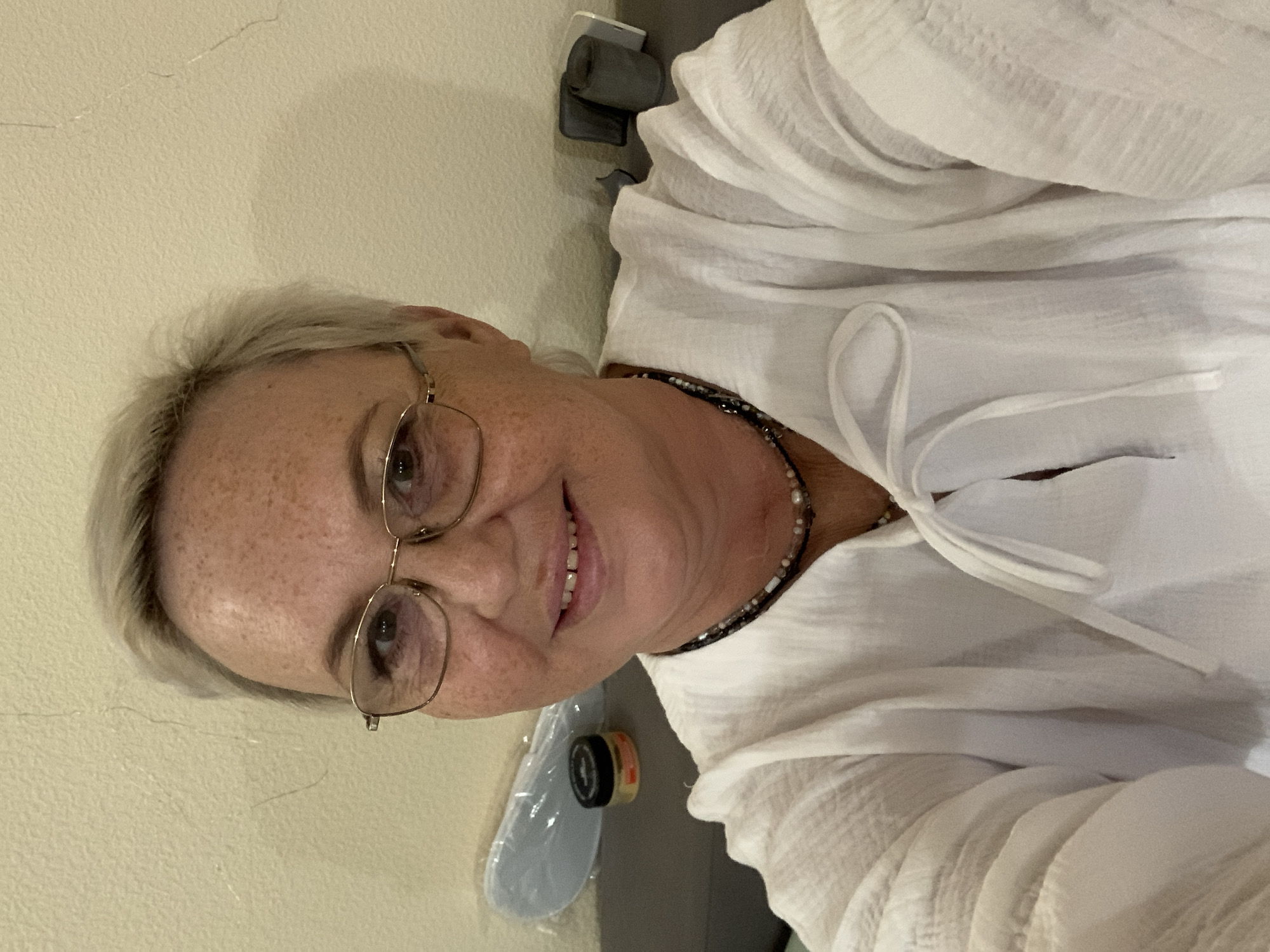 A woman with short, light-colored hair and glasses is taking a selfie indoors. She is wearing a white blouse with a tie at the neck and a beaded necklace. She has freckles on her face and is smiling. In the background, there is a wall, a gray surface, a small jar with a black lid, and a light blue object. There are also some dark, cylindrical objects on the gray surface to the right. The scene appears to be a room in a home or hotel. The location in the city of Chaikovsky, Russia is not discernible from the image.