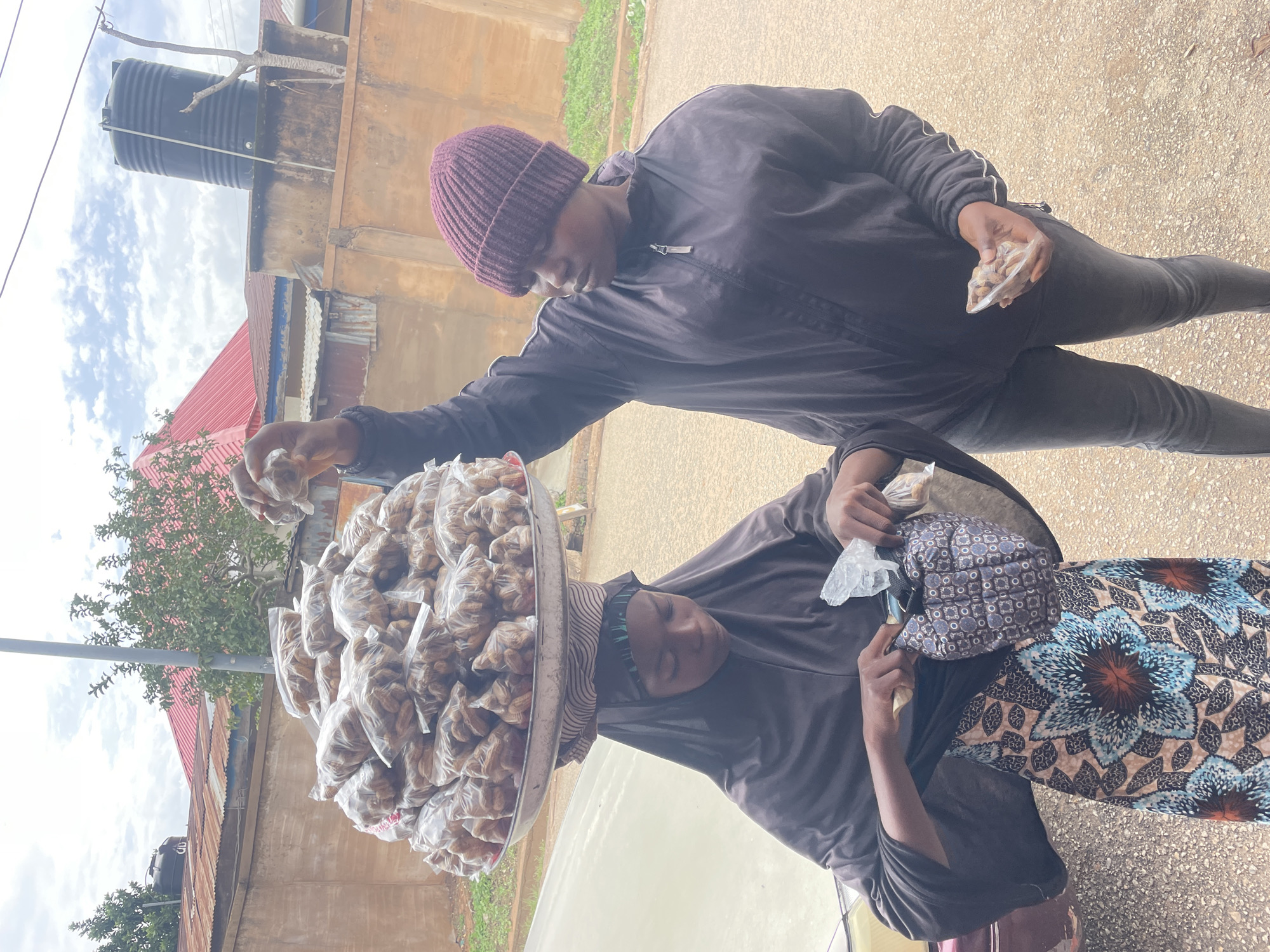 The image depicts an outdoor street scene in Jos, Nigeria, centered around a transaction between two women. One woman, the vendor, is seated or squatting, balancing a large, round metal tray filled with numerous small, clear plastic bags of snacks or nuts on her head. She wears a dark tunic, a vibrant patterned skirt with blue and brown floral motifs, and a striped headwrap. Her hands are engaged, possibly handing over goods or receiving payment.
Opposite her stands a customer, dressed in a dark hooded jacket, dark trousers, and a purple knitted beanie. She holds a small plastic bag containing similar snacks, indicating a recent or ongoing purchase. Both women are focused on their interaction.
The background reveals typical urban or semi-urban architecture, including buildings with a red corrugated iron roof and a multi-story structure with a black water tank prominently placed on its roof. Green trees and foliage are visible, and the ground appears to be paved. The bright, cloudy sky suggests it is daytime. The scene portrays a routine instance of street vending and local commerce. No visible text is present in the image.