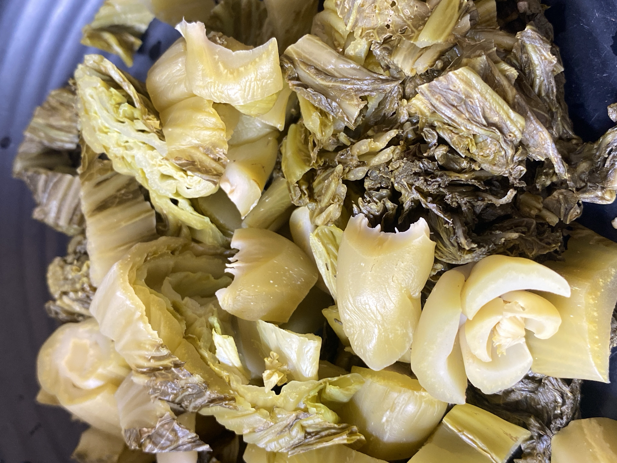 A close-up, overhead view shows a pile of pickled mustard greens in a dark bowl. The greens are a pale yellow-green and appear to be chopped into bite-sized pieces. Some pieces are still attached to stems, while others are separated leaves. The texture of the greens ranges from tender and slightly translucent to dried and wrinkled. The lighting is bright and even, highlighting the details of the food. The bowl appears to be made of metal and has a matte finish. The scene is entirely focused on the food, with no discernible background elements to indicate a specific location. There are no people or animals present in the image.