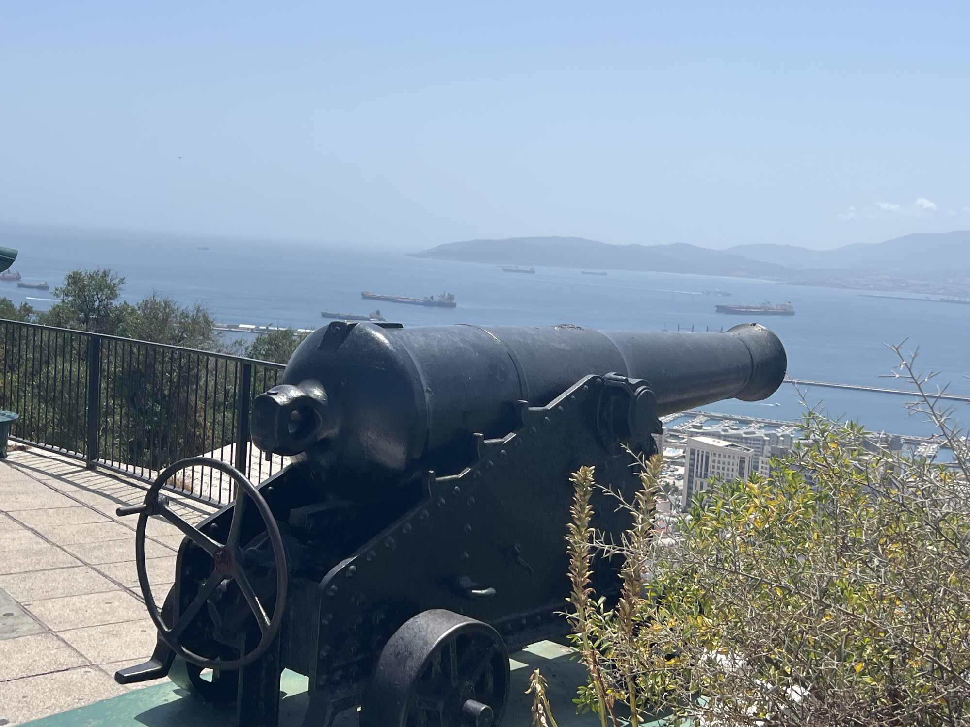 The image shows a historical cannon positioned on a paved viewing platform overlooking the Bay of Gibraltar. The cannon is black, made of metal, and appears to be in a static display, with a large wheel for aiming. Behind the cannon, a black metal fence runs along the edge of the platform, separating it from a steep, vegetated slope. In the background, the blue waters of the bay are dotted with numerous cargo ships and smaller vessels. The coastline of Spain or North Africa is visible in the distance, shrouded in a light haze. The scene is bathed in bright sunlight, suggesting a clear, warm day. The presence of the cannon, the panoramic view of the bay, and the distant coastline are characteristic of elevated vantage points in Gibraltar, such as the Great Siege Tunnels or the Upper Rock Nature Reserve, which offer historical context and strategic views of the strait.