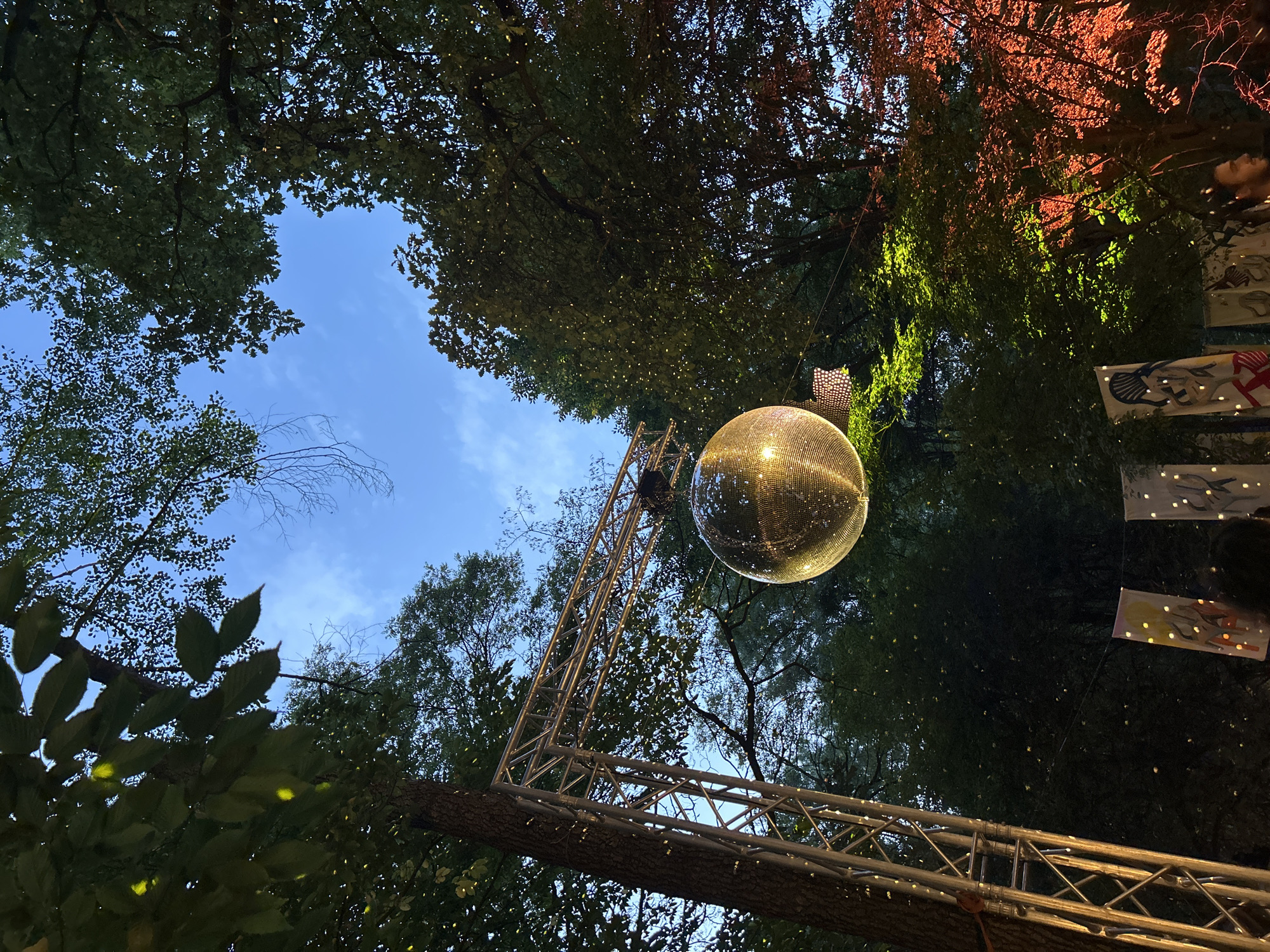 An outdoor setting at dusk features a large disco ball suspended from a metal truss structure. The disco ball is illuminated, reflecting light onto the surrounding trees. The trees are dense and leafy, with small, scattered lights twinkling within their branches, suggesting they are adorned with fairy lights. The sky above is a clear blue, with wisps of clouds. In the foreground, several hanging banners with illustrated designs are visible, and a few people are partially seen at the bottom of the frame. The scene appears to be a festive gathering or outdoor event. The location is Unknown, Unknown.