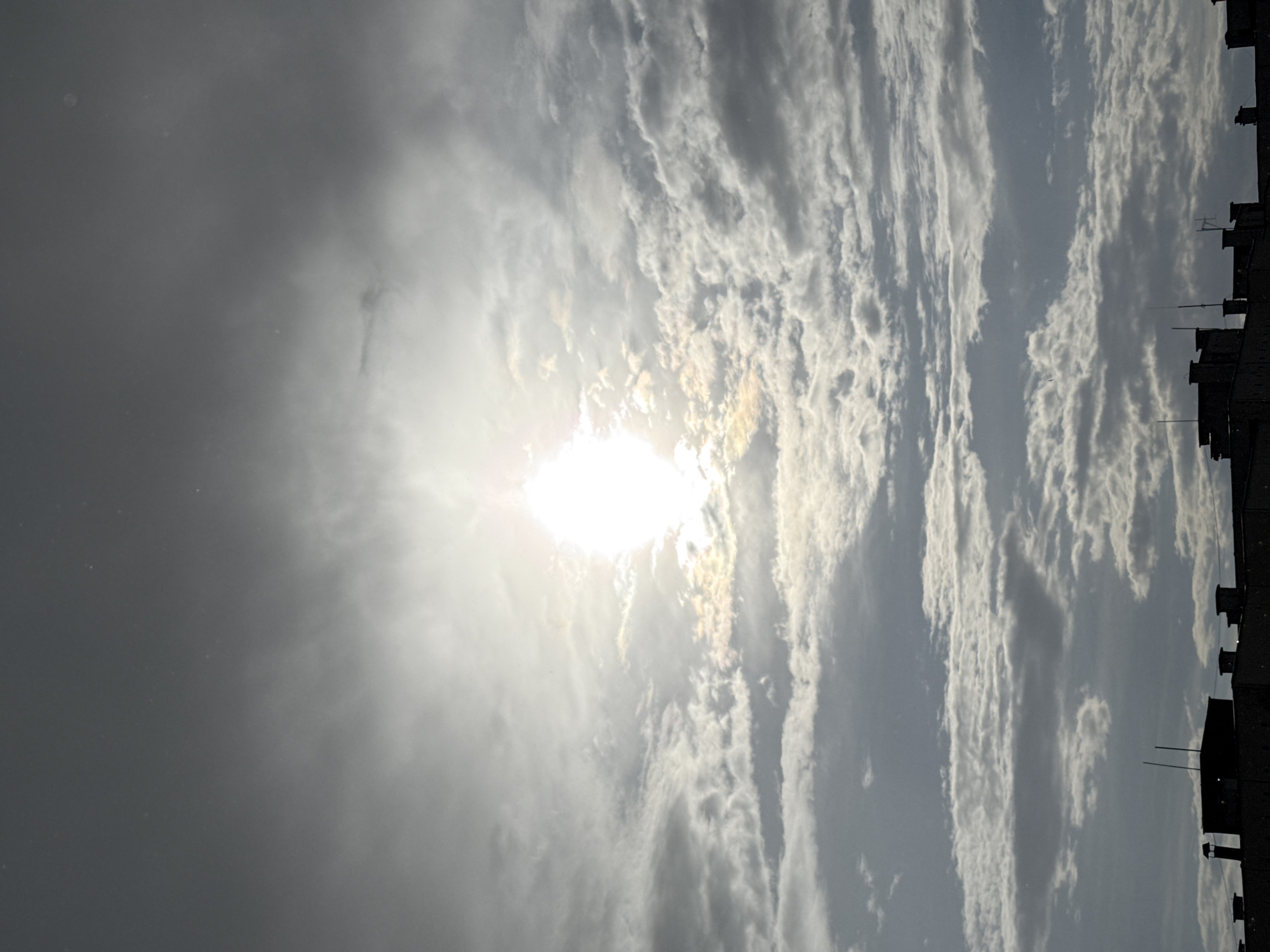 The image shows a daytime sky above Pleven, Bulgaria. The sun is visible, obscured by thin clouds. The sky is a mix of gray and white clouds. The silhouette of a building rooftop with chimneys and antennas is seen along the right edge of the frame.