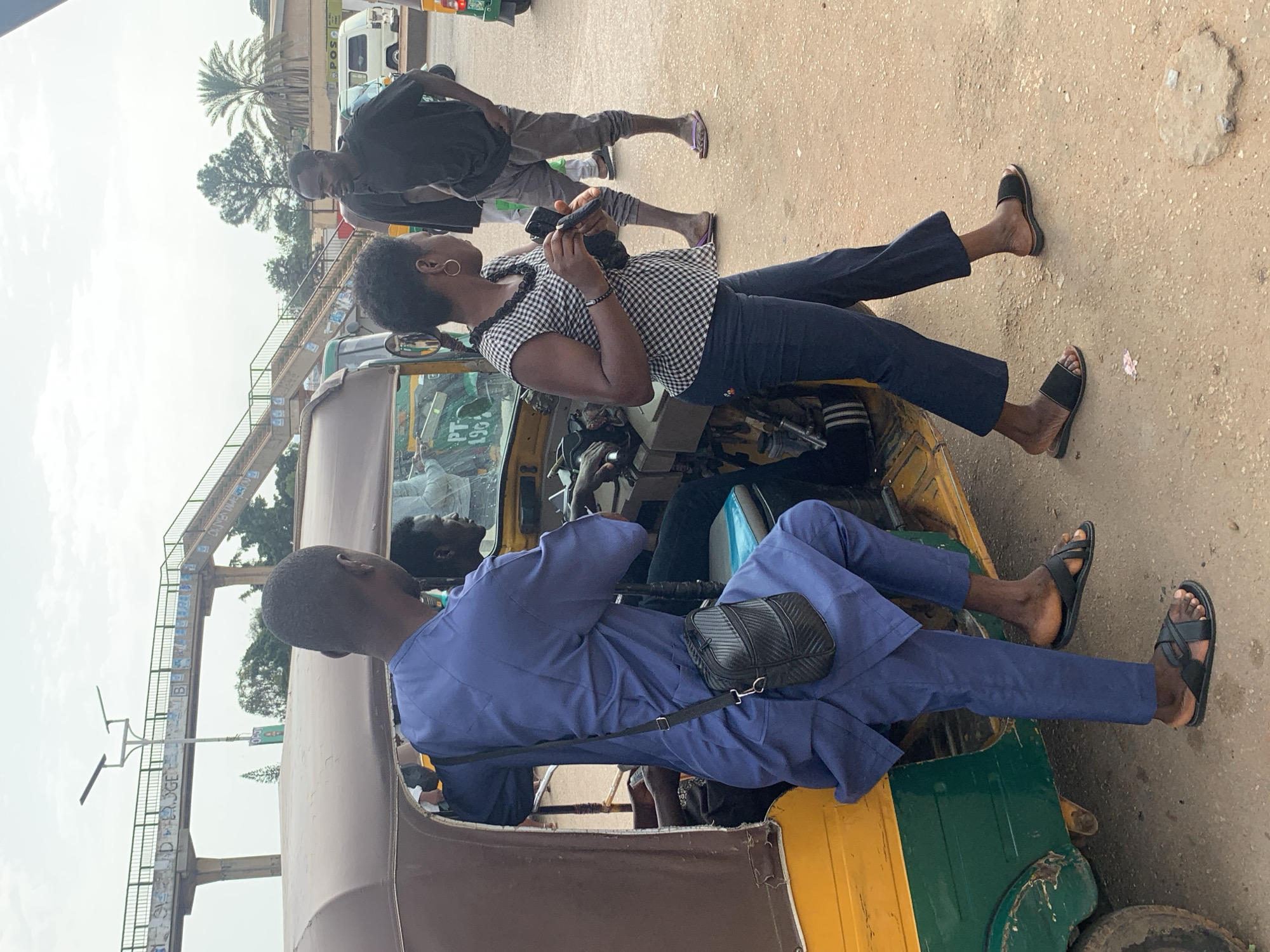 The image depicts a street scene in Gura Top, Nigeria, under an overcast sky. In the foreground, a yellow and green auto-rickshaw is parked. A man, dressed in a flowing blue traditional garment and black sandals, is partially visible entering the auto-rickshaw from the side. He carries a black crossbody bag. Next to him, a woman wearing a black and white checkered blouse and dark pants, adorned with hoop earrings, is holding a mobile phone. She is looking towards the auto-rickshaw. Further in the background, a man in a black t-shirt and gray pants stands, his gaze directed away from the camera. In the middle ground, a pedestrian bridge with graffiti runs across the scene, supported by concrete pillars. Trees and buildings are visible behind the bridge, along with other vehicles including a white van. The ground is a mix of dirt and gravel.