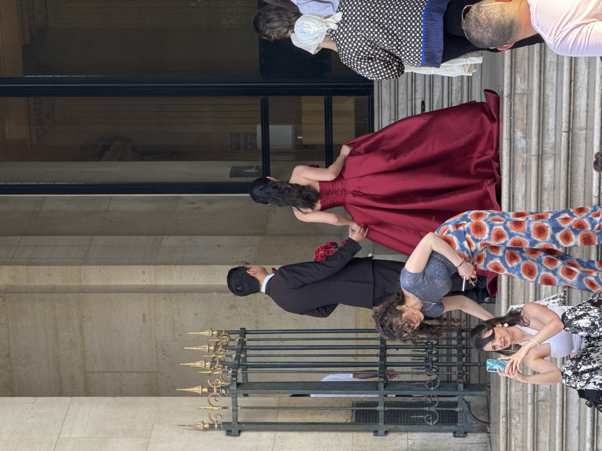 A wedding ceremony is taking place at the Petit Palais in Paris, France. A groom in a black suit and a bride in a burgundy dress are exiting the building, holding hands. Two women are taking photos of the couple with their cell phones. Several people can be seen at the top of the stairs. The building has ornate architecture.