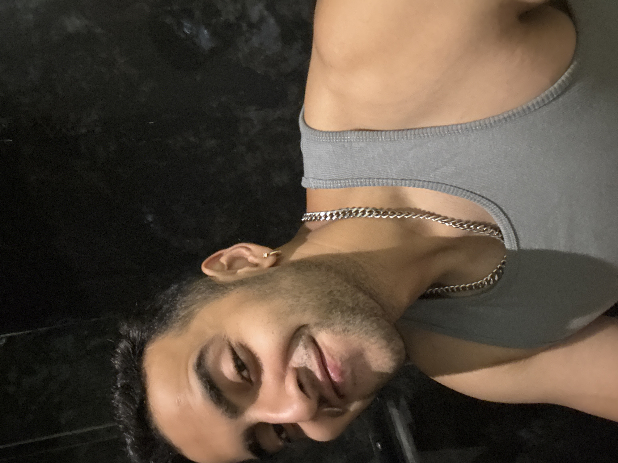 This close-up portrait, taken in Kolkata, India, features a young adult male depicted from the chest up, with the image rotated ninety degrees clockwise. The man looks directly at the viewer with a subtle, pleasant smile, suggesting a confident or friendly demeanor.
He has dark hair, styled short, and visible stubble or a closely trimmed beard on his face. A small, golden hoop earring is visible in his left ear. He is wearing a light grey, ribbed tank top that conforms to his torso. Around his neck, he sports a silver-colored curb chain necklace, prominent against his skin and the fabric of his shirt.
The background is dark and appears to be a plain, possibly textured wall or surface, which provides a simple, undiscernible setting. The lighting highlights the subject, suggesting an indoor environment or a photo taken in low-light conditions, likely in the evening or at night. There are no other discernible objects or activities taking place, and no text is visible in the image.