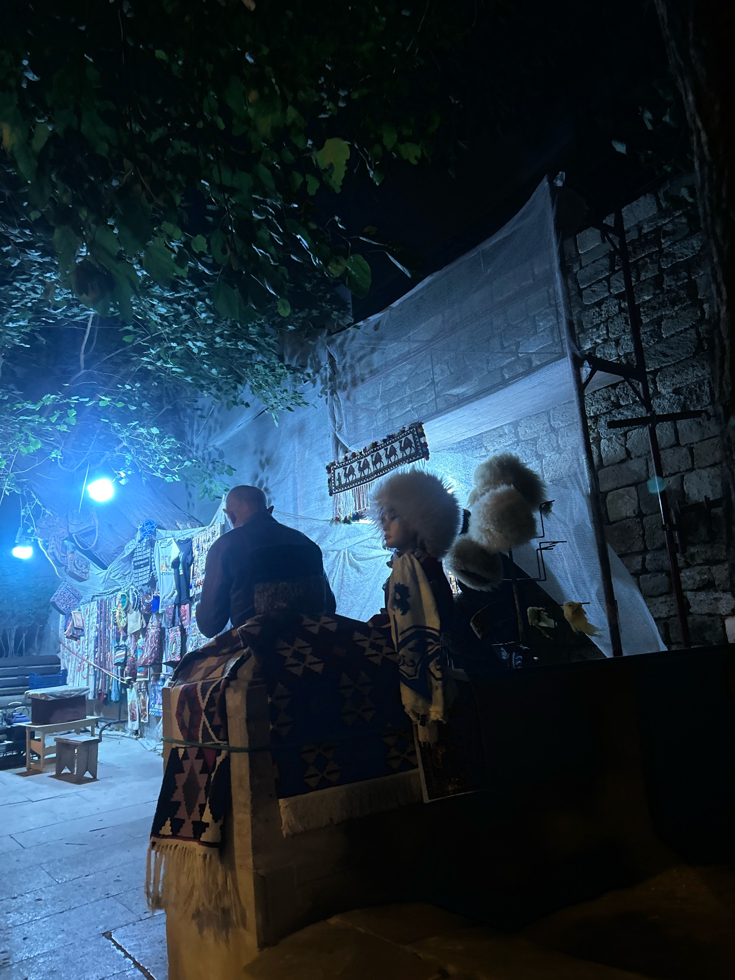 The image depicts an outdoor marketplace at night, possibly in Sabail Raion, Azerbaijan, illuminated by blue and white lights. In the foreground, two figures are partially visible. One individual, seen from the back, is wearing a dark jacket and is seated on what appears to be a decorated surface with intricate patterns. In front of them, to the right, is another figure wearing a distinctive white, fluffy hat and a patterned garment. Behind these figures, a stall displays various textiles, bags, and trinkets, with some items hanging and others folded. A white sheet or tarp serves as a backdrop for the stall, contrasting with a rough stone wall on the right side of the image. Overhanging from the top left are dark green leaves, suggesting the presence of trees. The ground appears to be paved.