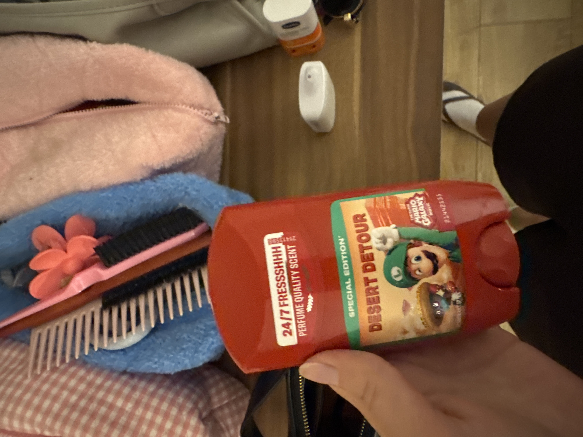 The image shows a person holding a red deodorant stick with a "Desert Detour" label featuring characters from "The Super Mario Galaxy Movie." In the background, a pink fuzzy bag and a blue towel are visible, along with various grooming items like combs and brushes. The scene appears to be indoors, possibly in a bathroom or bedroom, with a wooden surface in the foreground. There is no visible indication of the city of Aksu, Turkey in the image.