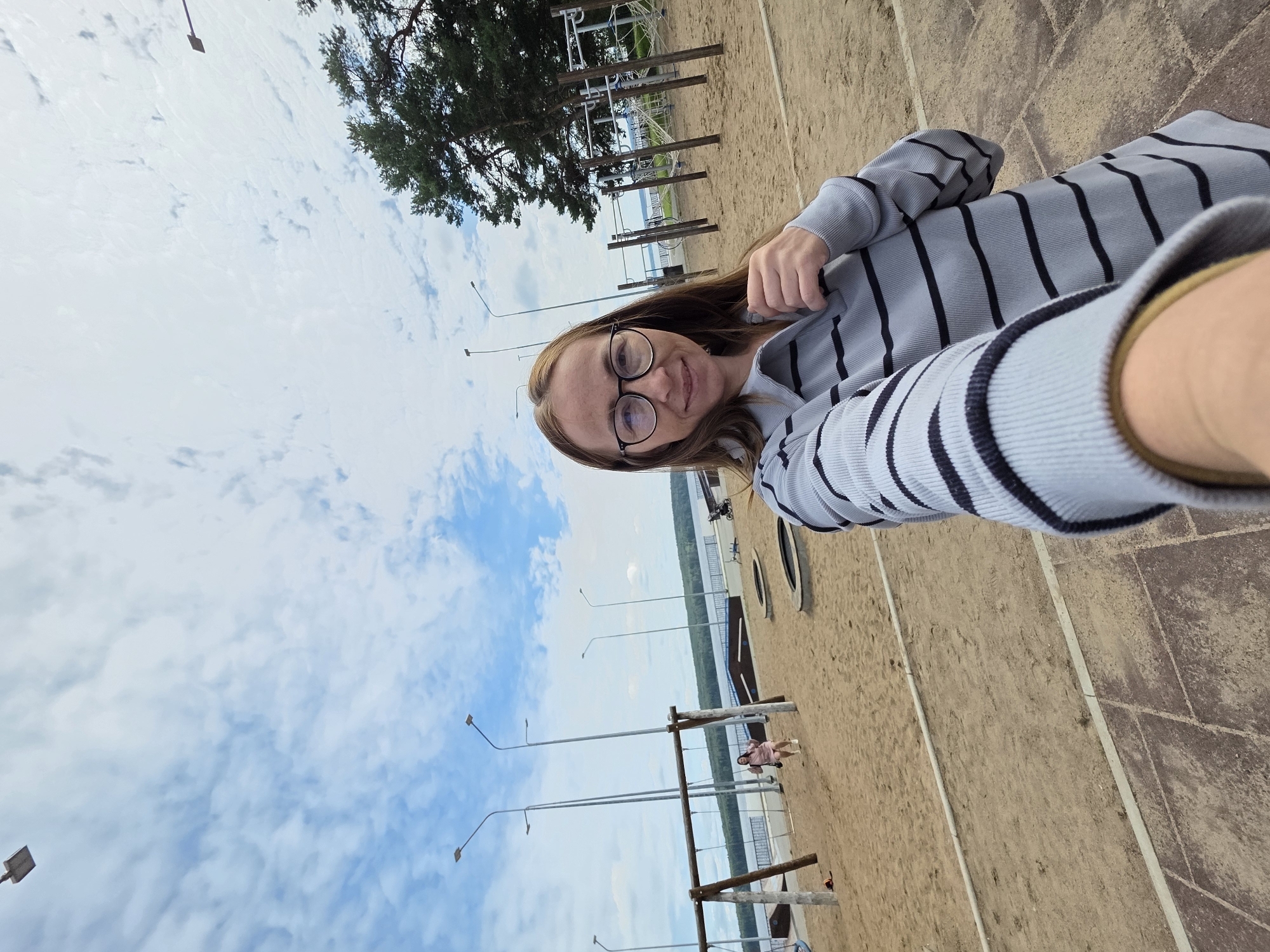 The image shows a selfie taken outdoors on a sandy beach or playground area with a body of water and trees in the background under a cloudy sky. In the foreground, a woman wearing glasses and a striped long-sleeved shirt is visible from the chest up. She is smiling at the camera and holding her arm out to take the picture. In the middle ground, there is a playground with swings and climbing equipment, and a young girl on a swing. The background features a wide expanse of water, a tree line, and several streetlights. This scene is located at the beach on the Kama River embankment in Chaikovsky, Russia, a city known for its scenic waterfront.