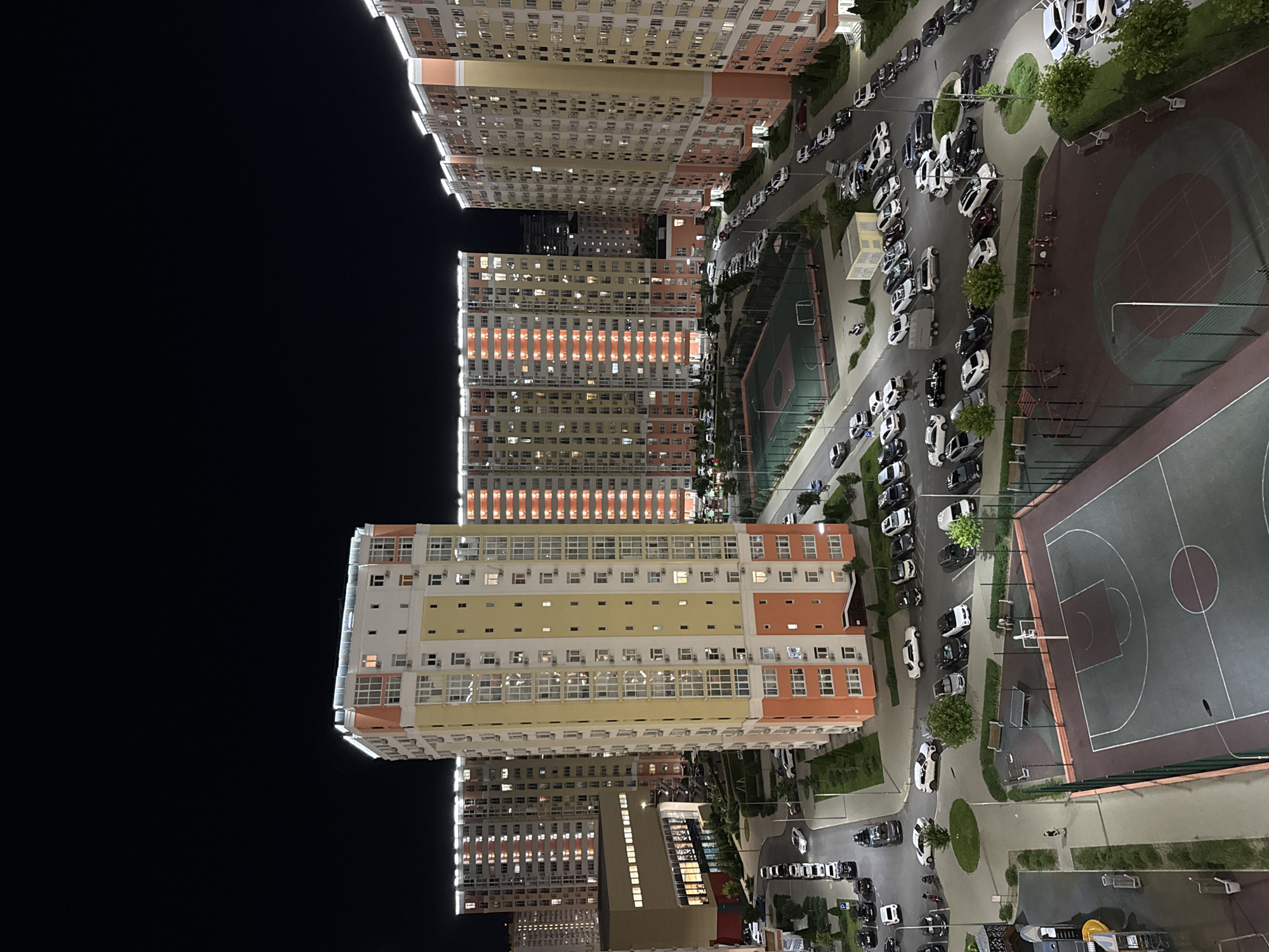 The image shows a night-time aerial view of a residential complex in Anapa, Russia. Several tall, multi-story apartment buildings with illuminated windows are prominent. The buildings are primarily cream and orange in color and feature balconies. In front of the buildings, a network of roads and parking lots are filled with parked cars. There are also recreational areas visible, including a basketball court with markings and a hoop, as well as an outdoor gym with various exercise equipment. A few people are present in the recreational areas. The overall scene depicts a modern urban residential development at night.
