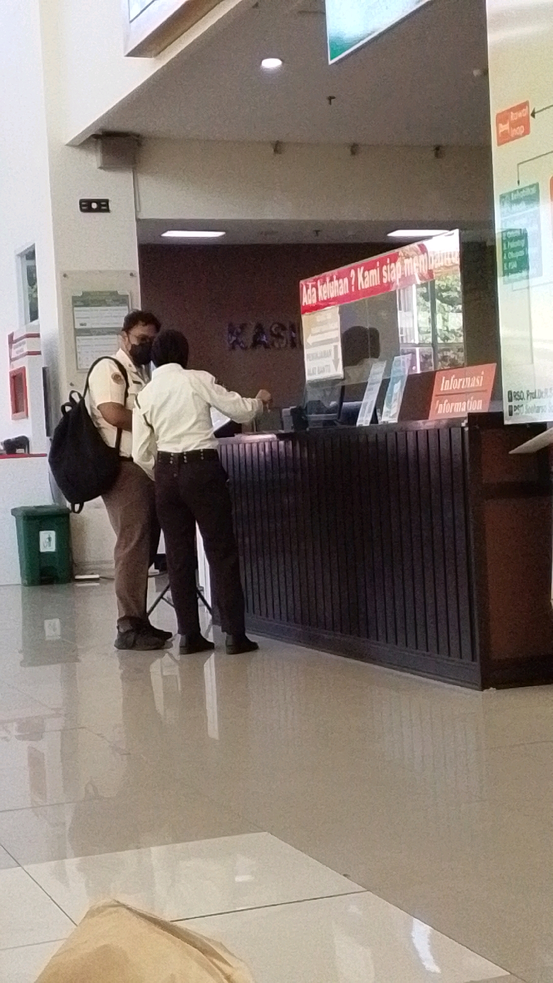 In a brightly lit indoor environment, possibly a hospital or public service building in Pabelan, Indonesia, two individuals are interacting at a counter. The person on the left, wearing a light brown uniform with a dark backpack slung over their shoulder and a face mask, appears to be engaging with the person behind the counter. The person behind the counter, dressed in a white long-sleeved shirt and dark pants, is also wearing a face mask and is holding a pen, seemingly attending to a transaction or providing information. Behind them, a large wooden counter with vertical slats is visible, with various signs and possibly a computer monitor on top. One sign reads "Informasi Information," and another partially visible sign above the counter says "KASI." To the left of the counter, a green trash can is present. The floor is polished and reflective, showing the reflections of the people and objects. In the background, a large sign with Indonesian text, including "RAWAT INAP," is displayed, indicating this is likely a healthcare facility. The overall scene suggests a typical day at a service counter where individuals are receiving assistance or conducting administrative tasks.