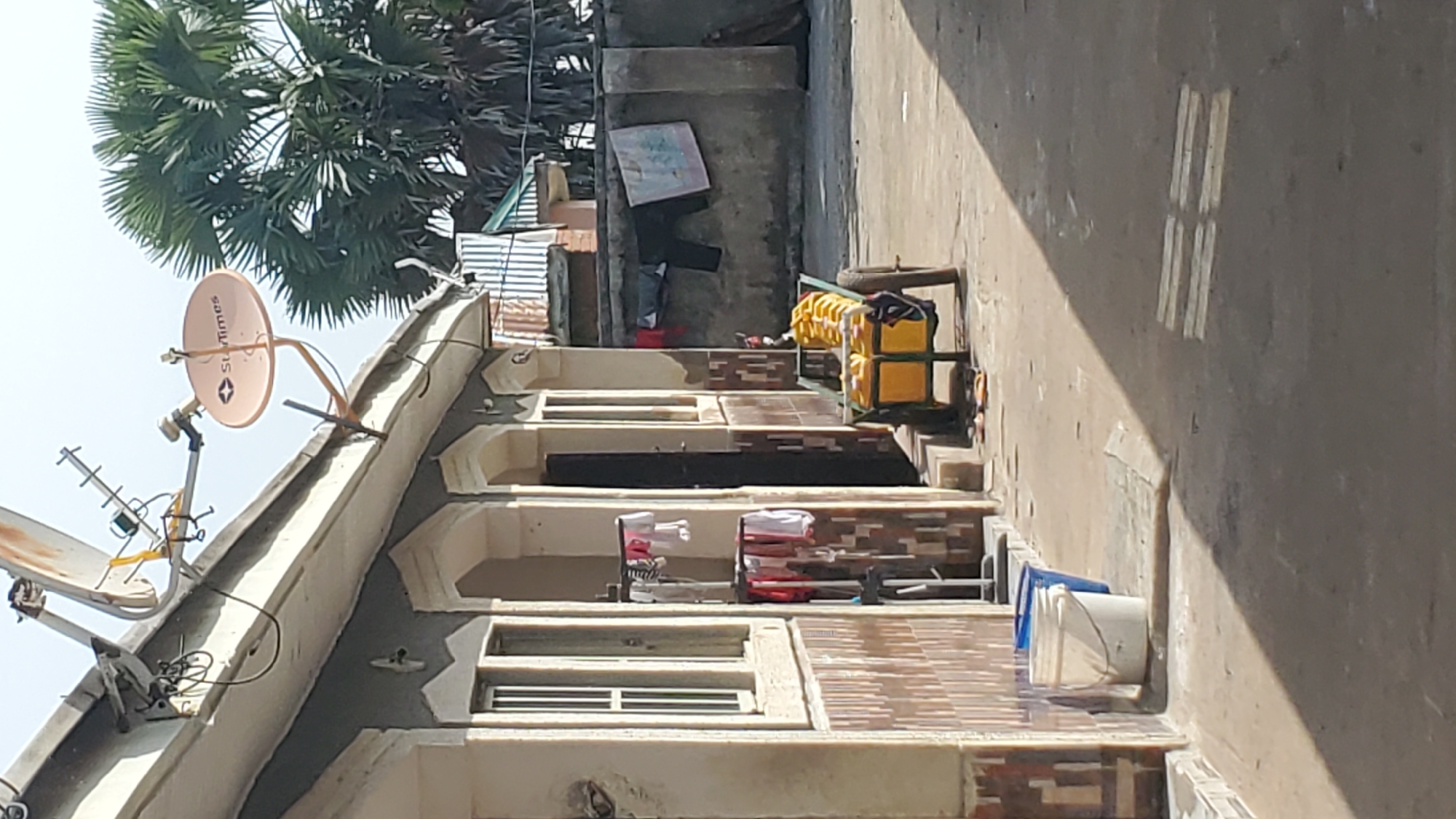 The image captures an outdoor scene in Jalingo, Nigeria, with a focus on a residential building and its surroundings. The building, partially visible, features a light-colored exterior with decorative tiled sections on its lower facade. Several windows with white frames and grates are present, suggesting security measures. Above the building, multiple satellite dishes and an antenna are mounted on the roof, indicating access to broadcast services.
In the foreground, a paved pathway is evident, with shadows cast by an unseen structure, creating a stark contrast. To the left, two buckets, one white and one blue, are placed near the building. Further into the scene, a yellow handcart is loaded with several yellow plastic containers, possibly for water or other liquids. Laundry hangs on a clothesline attached to a vertical pole, with various garments visible, including shirts and undergarments. In the background, a concrete wall runs horizontally, and laundry is also hung on it, including a pair of dark trousers and a patterned cloth. A large palm tree with spiky fronds stands prominently behind the wall, adding a tropical element to the environment. The ground appears to be a mix of compacted earth and concrete. The overall impression is of a typical residential courtyard in an urban Nigerian setting, with everyday activities like laundry and water storage in plain sight.