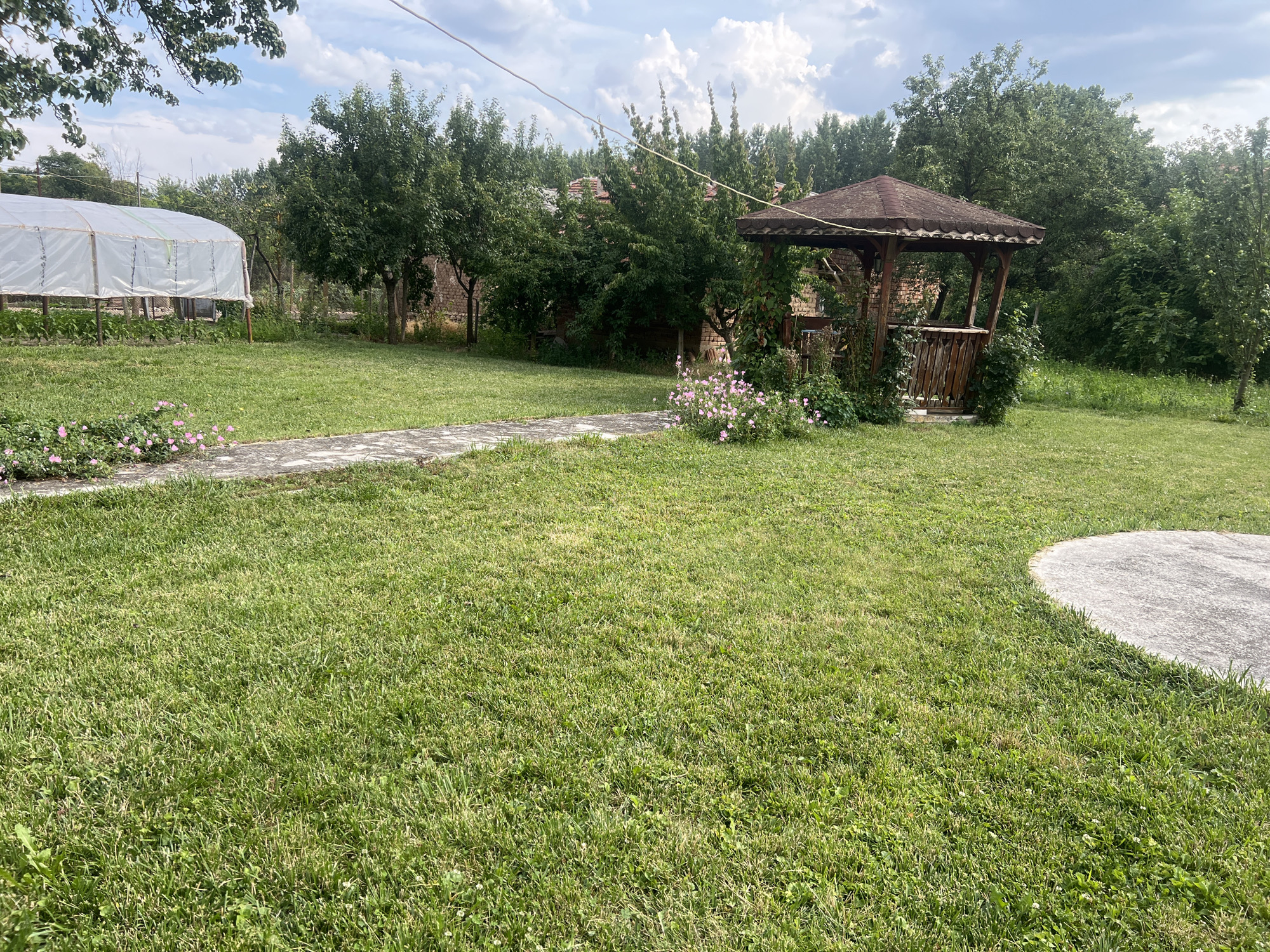 The image depicts a backyard scene with a wooden gazebo in the center, surrounded by lush green grass and trees. A paved walkway curves through the lawn, leading towards the gazebo. To the left, a greenhouse structure is visible, covered with white plastic sheeting. In the background, several houses with tiled roofs and brick walls are interspersed with dense foliage. The sky is partly cloudy. The location is identified as Ohrid, Bulgaria. There are no people or animals visible in the image.