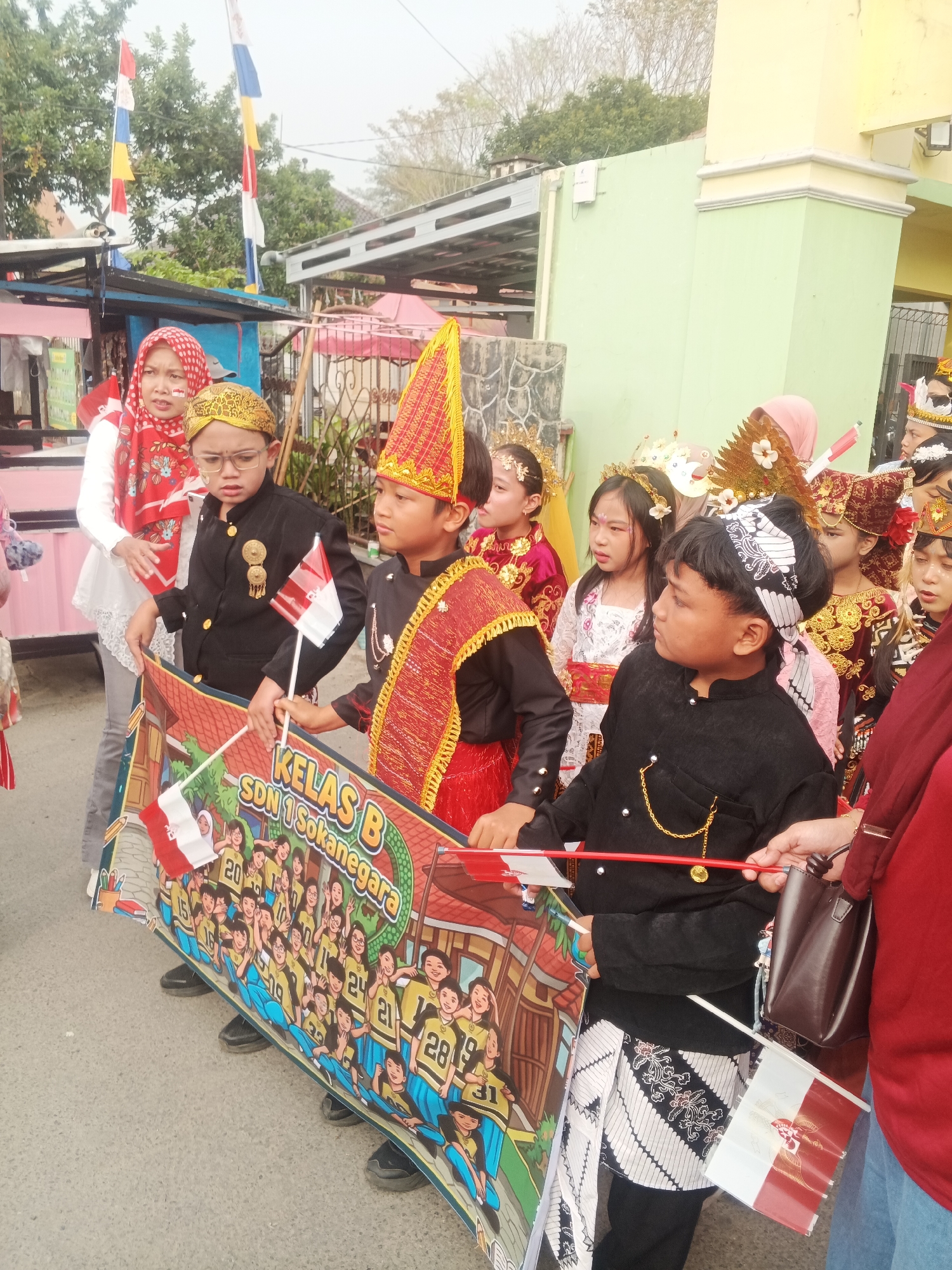 A group of children in traditional attire are standing in a procession in Sokanegara, Indonesia. The children, some wearing elaborate headdresses and sashes, are holding a banner that reads "KELAS B SDN 1 SOKANEGARA". The banner depicts cartoon-like images of other children in uniform. Two children in the foreground are holding small Indonesian flags. Behind the children, a few adults are visible, including a woman in a red headscarf and a woman in a red shirt carrying a dark brown purse. The background shows buildings and trees, suggesting an outdoor urban or semi-urban setting. The overall scene appears festive, likely part of a school event or parade.