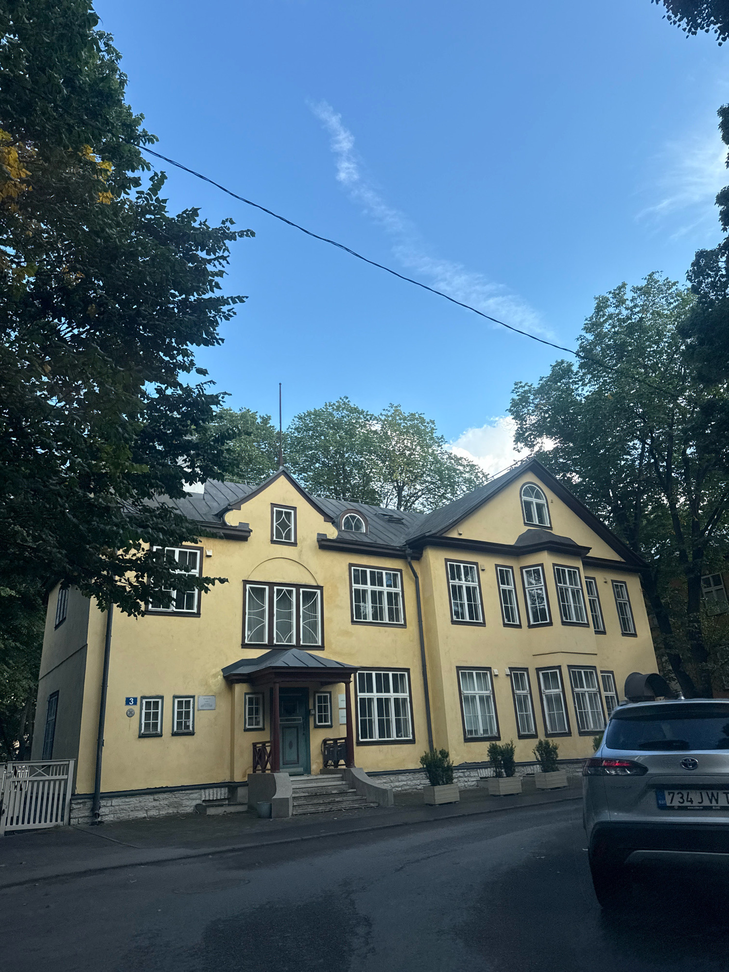 The image captures a light yellow, two-story building with a dark roof, situated on a street in Tallinn, Estonia. The building features numerous white-framed windows, some with decorative grilles, and a prominent entrance with a covered porch. Trees with green foliage frame the building on both sides, and a clear blue sky with a wispy white cloud is visible above. A silver SUV with a license plate reading "734 JWT" is parked on the right side of the road in front of the building. The street is paved, and a white picket fence is visible to the left of the building. A blue house number "3" is displayed on the facade near the entrance.
