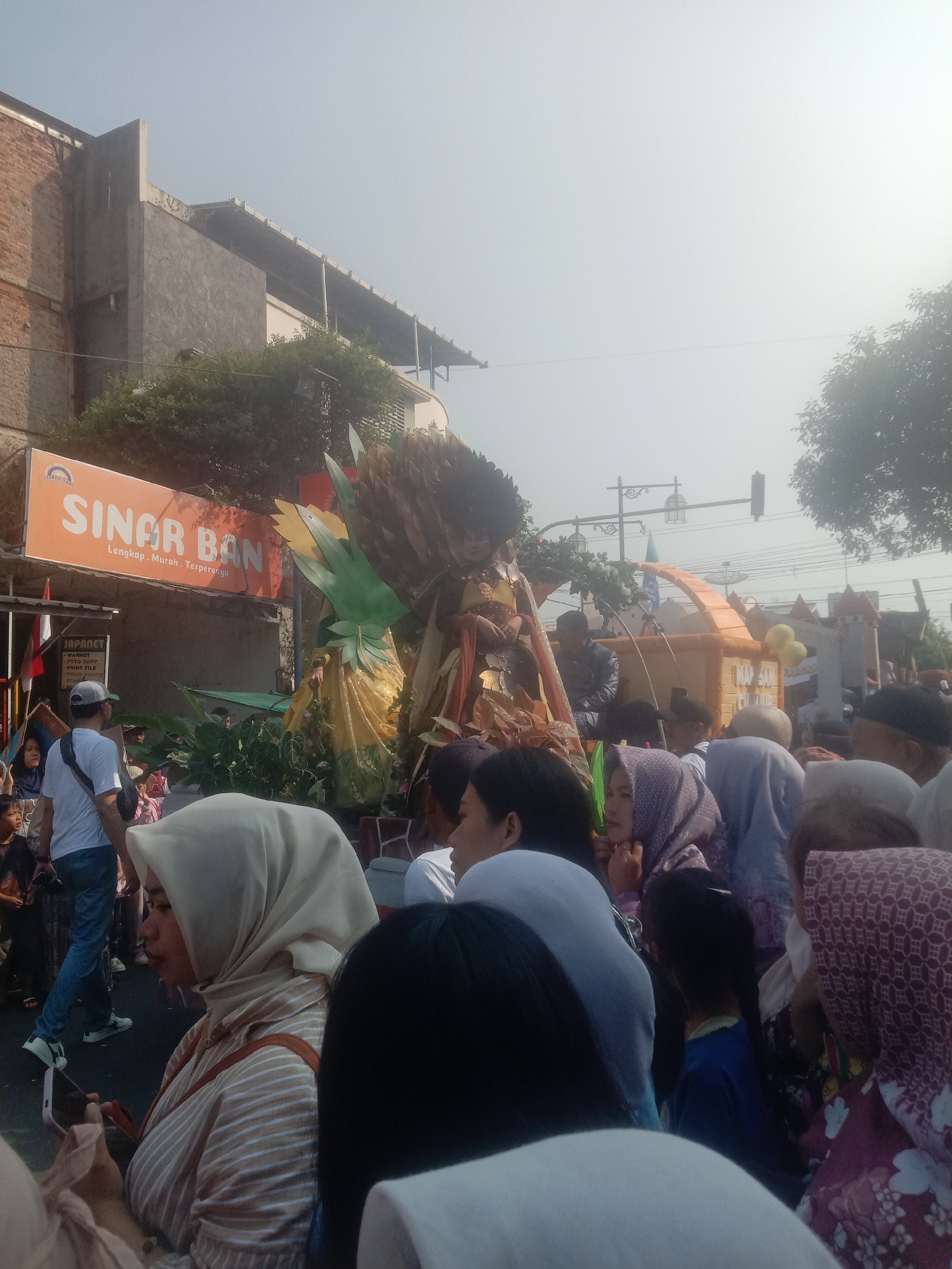 The image captures a street scene in Cianjur, Indonesia, likely during a parade or festival. In the foreground, a crowd of people are visible, many wearing headscarves. Some are looking towards a decorated float in the middle ground. The float features a figure adorned in a golden outfit, surrounded by large, leafy green decorations and what appear to be golden wheat stalks. Behind the float, a bright orange sign reads "SINAR BAN" in white lettering, with smaller text below in Indonesian. Further in the background, buildings and trees are visible, and the sky is hazy. The overall atmosphere suggests a public event with a festive and crowded environment.