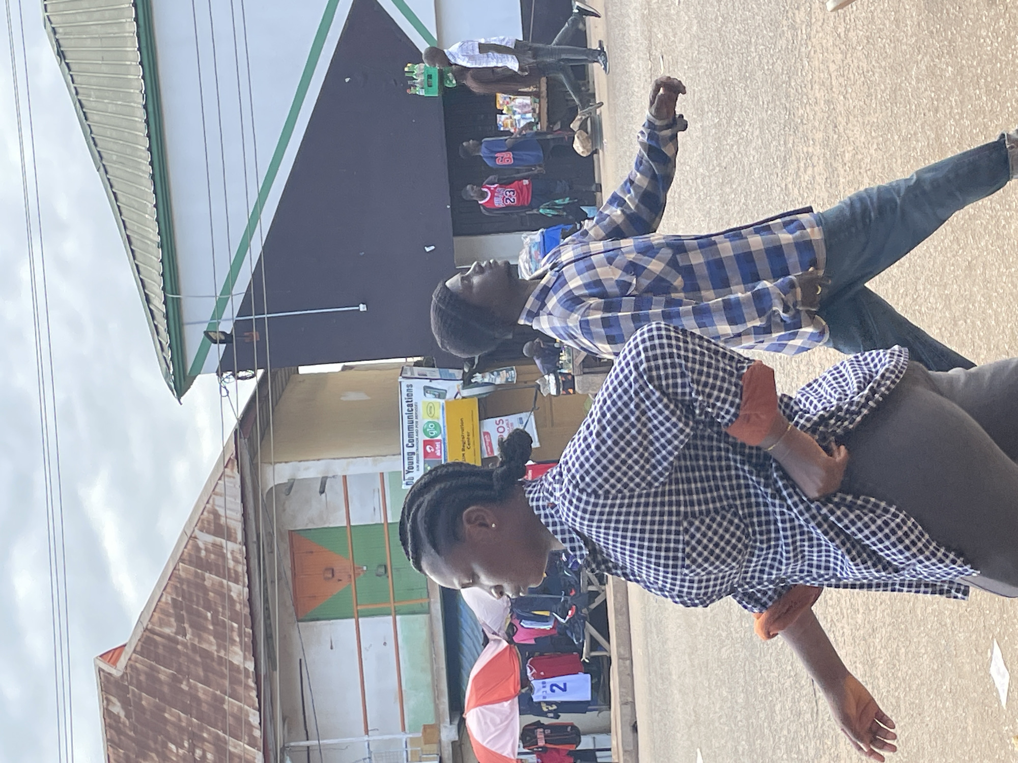 The image depicts an outdoor scene in Bukuru, Nigeria, with several people visible. In the foreground, two individuals are the primary subjects, both wearing checkered shirts. The person on the right is mostly obscured, but their arm and hand are visible, suggesting movement. The person on the left, a woman, is looking down and to the side, with her hair in braids. She is wearing a blue and white checkered shirt. Behind them, other people are seen walking or standing. One individual in the background is wearing a red basketball jersey with the number 23.
The setting appears to be a street or market area. There are buildings with signage, including a dark facade with a green stripe and what looks like a corrugated metal roof on a smaller structure to the left. The ground is paved. The lighting suggests it is daytime, possibly overcast, given the diffuse light.
There is visible text on signage in the background. One sign reads "Young Communications" with logos for "Airtel" and "Glo". Another sign has "SOS". An orange and pink umbrella is also visible, partially open. The overall atmosphere suggests a casual, everyday scene in a bustling urban environment.